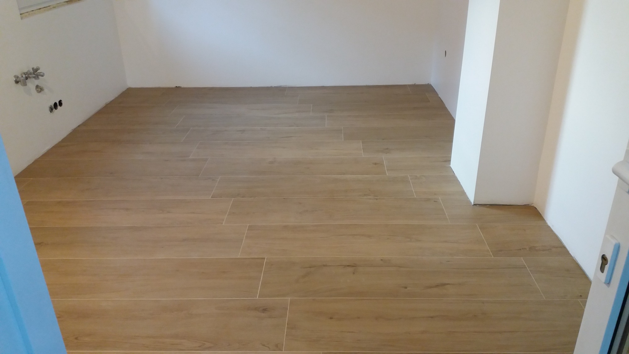 Ukończona podłoga z płytek imitujących drewno w jasnym odcieniu, ułożonych w nowym, minimalistycznym wnętrzu z białymi ścianami i widocznymi przyłączami hydraulicznymi.