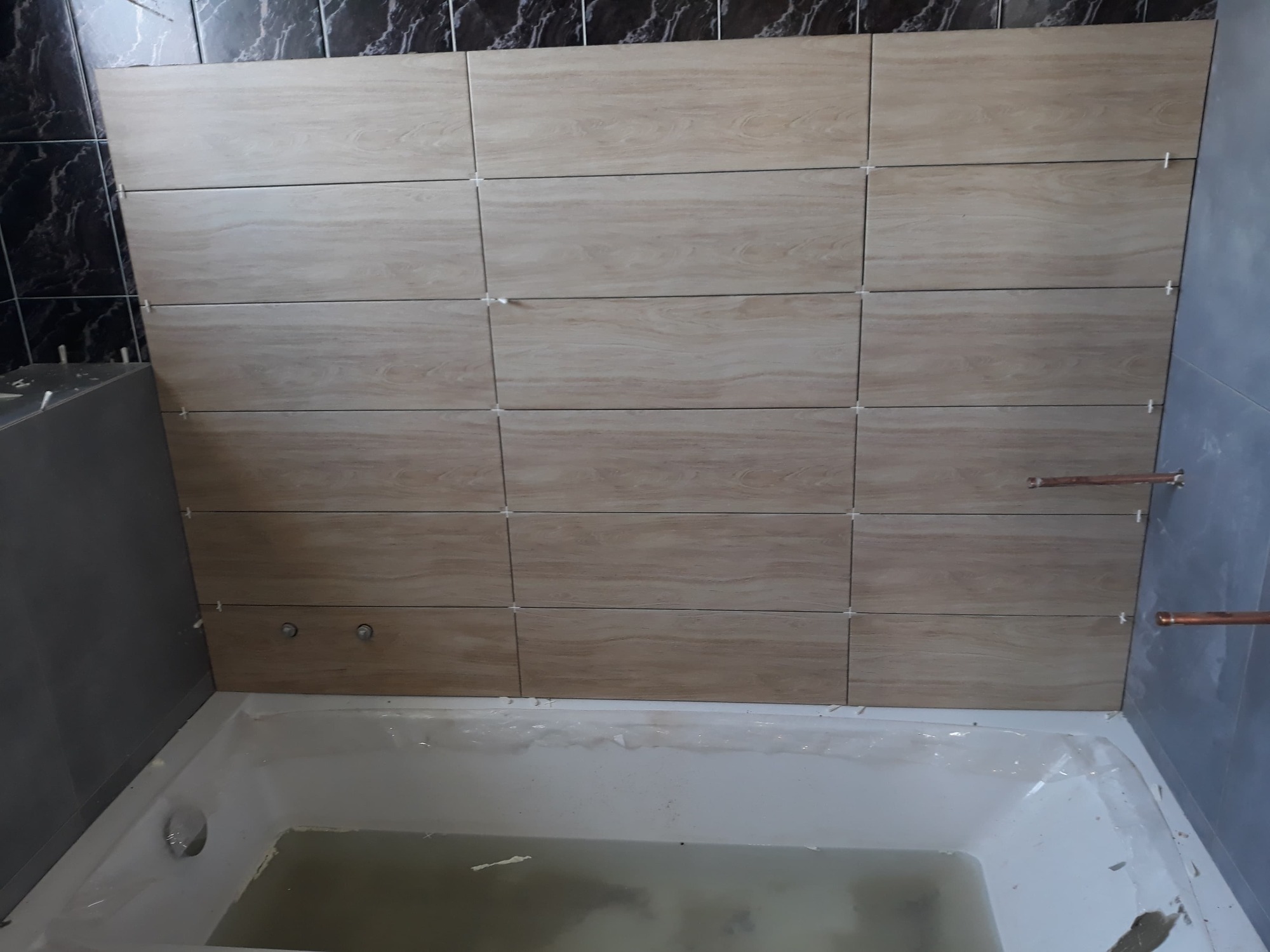 Ściana nad wanną w trakcie wykańczania, widoczne płytki imitujące drewno z krzyżykami dystansowymi, rury miedziane wystające ze ściany, wanna częściowo zakryta folią ochronną.