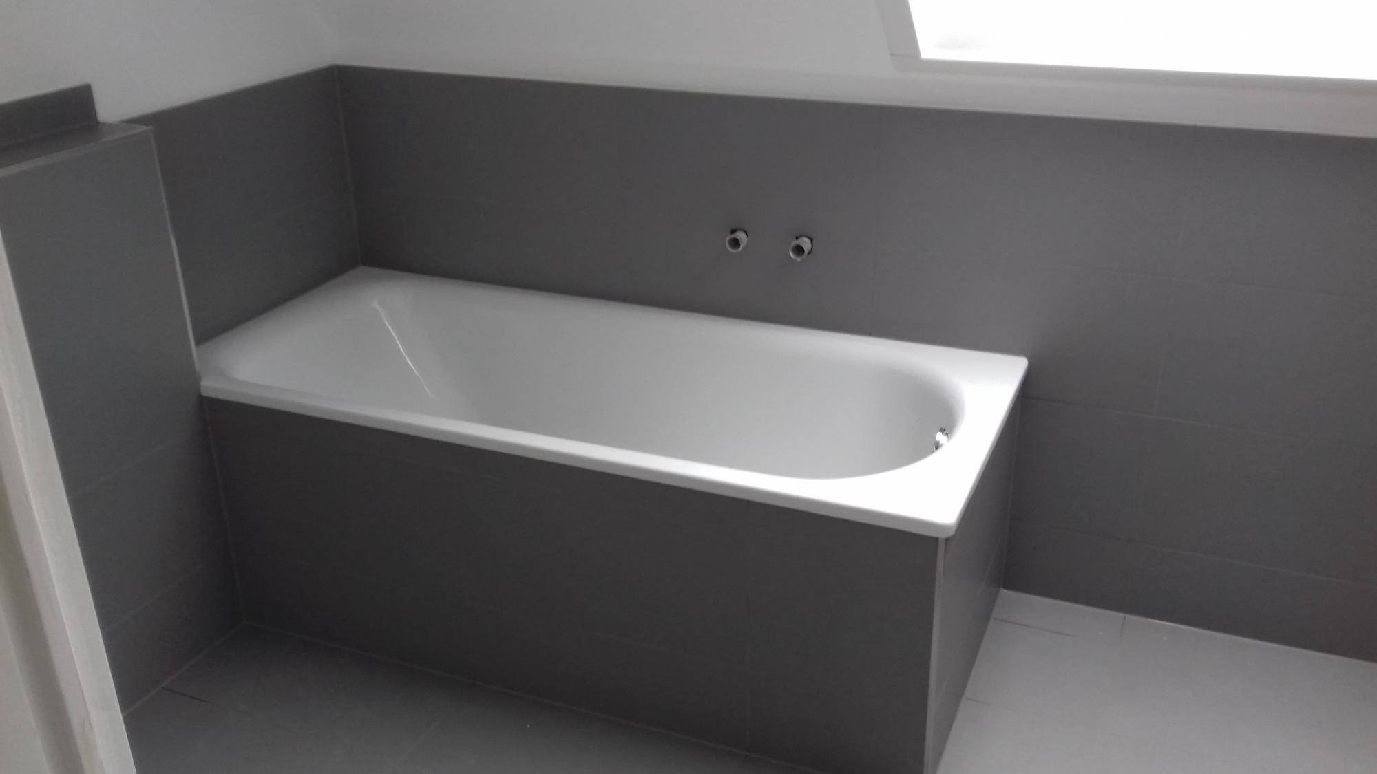 Wykończona łazienka z wanną obudowaną szarymi płytkami i szarą glazurą na ścianach, widoczne wystające rury do baterii.