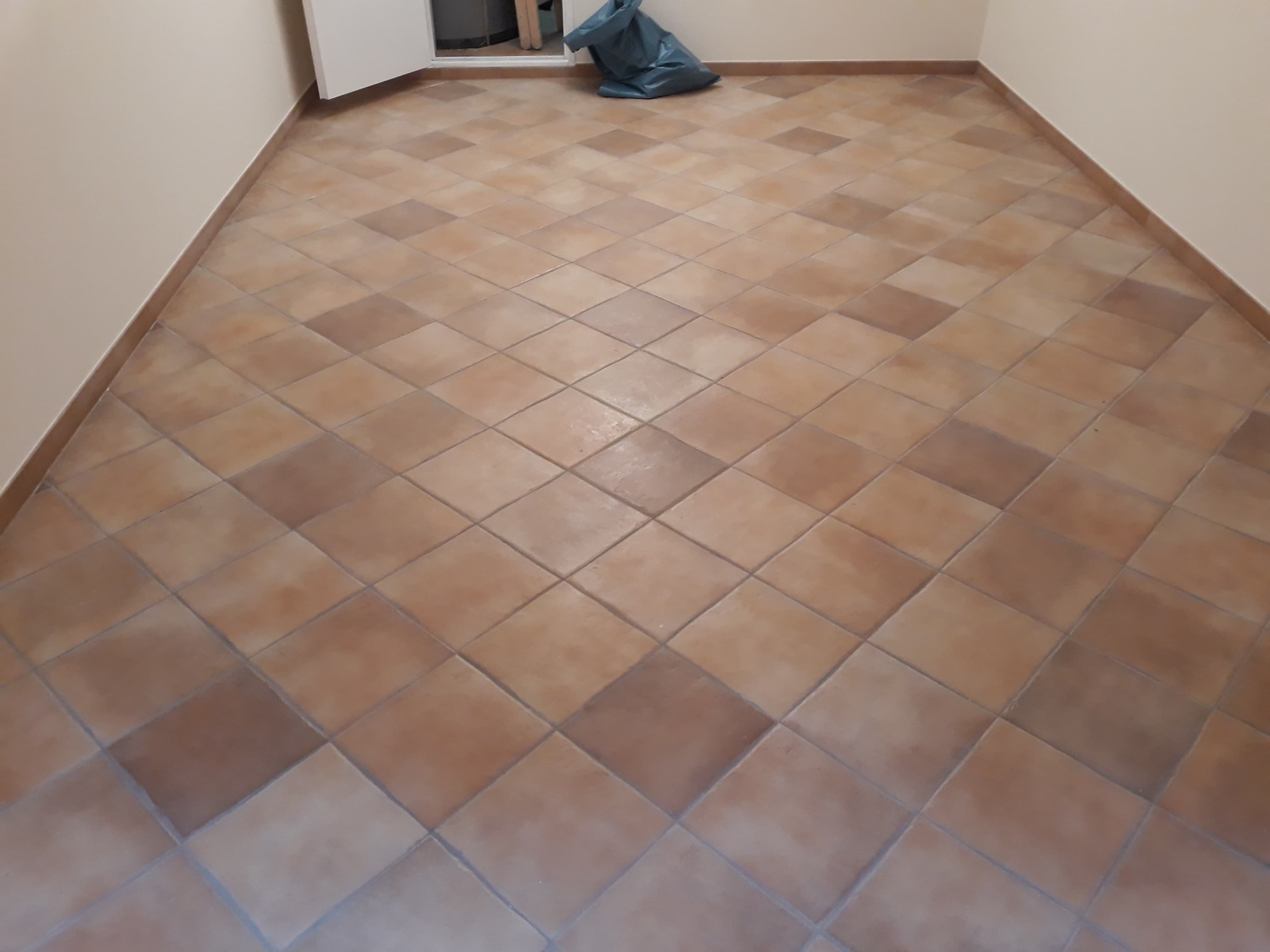 Ukończona podłoga wyłożona kwadratowymi, terakotowymi płytkami ułożonymi w karo, widoczny worek na śmieci w rogu pokoju.