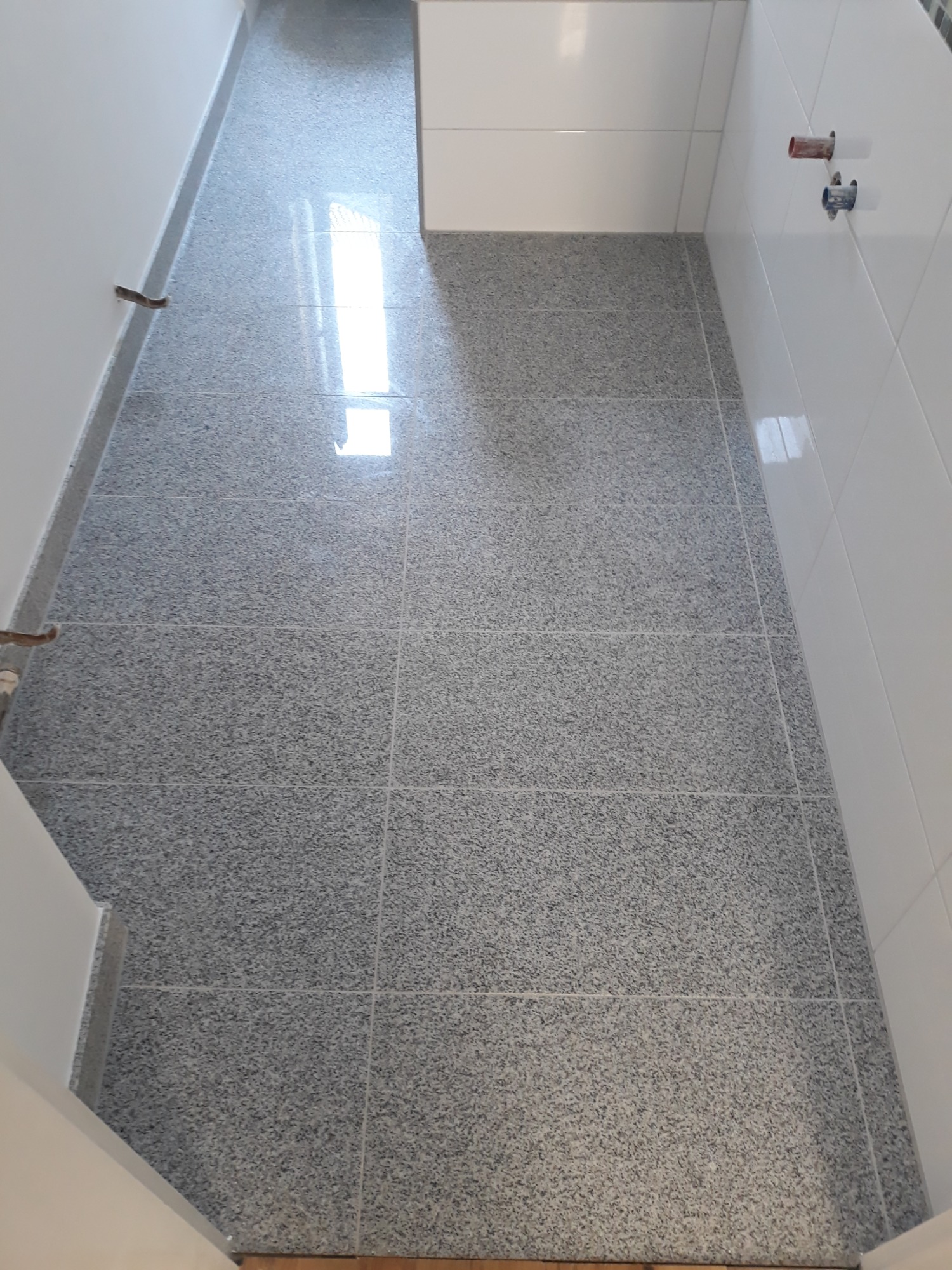 Szare, nakrapiane płytki podłogowe ułożone w łazience, widoczne białe ściany z fragmentem glazury i wystające rury instalacji wodnej.