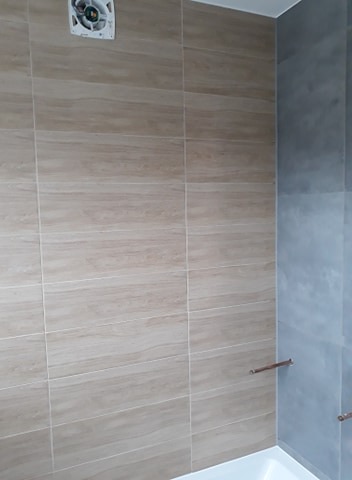 Wykończona łazienka z beżowymi płytkami imitującymi drewno na ścianie, szarymi płytkami w rogu, widoczny wentylator sufitowy i fragment wanny.