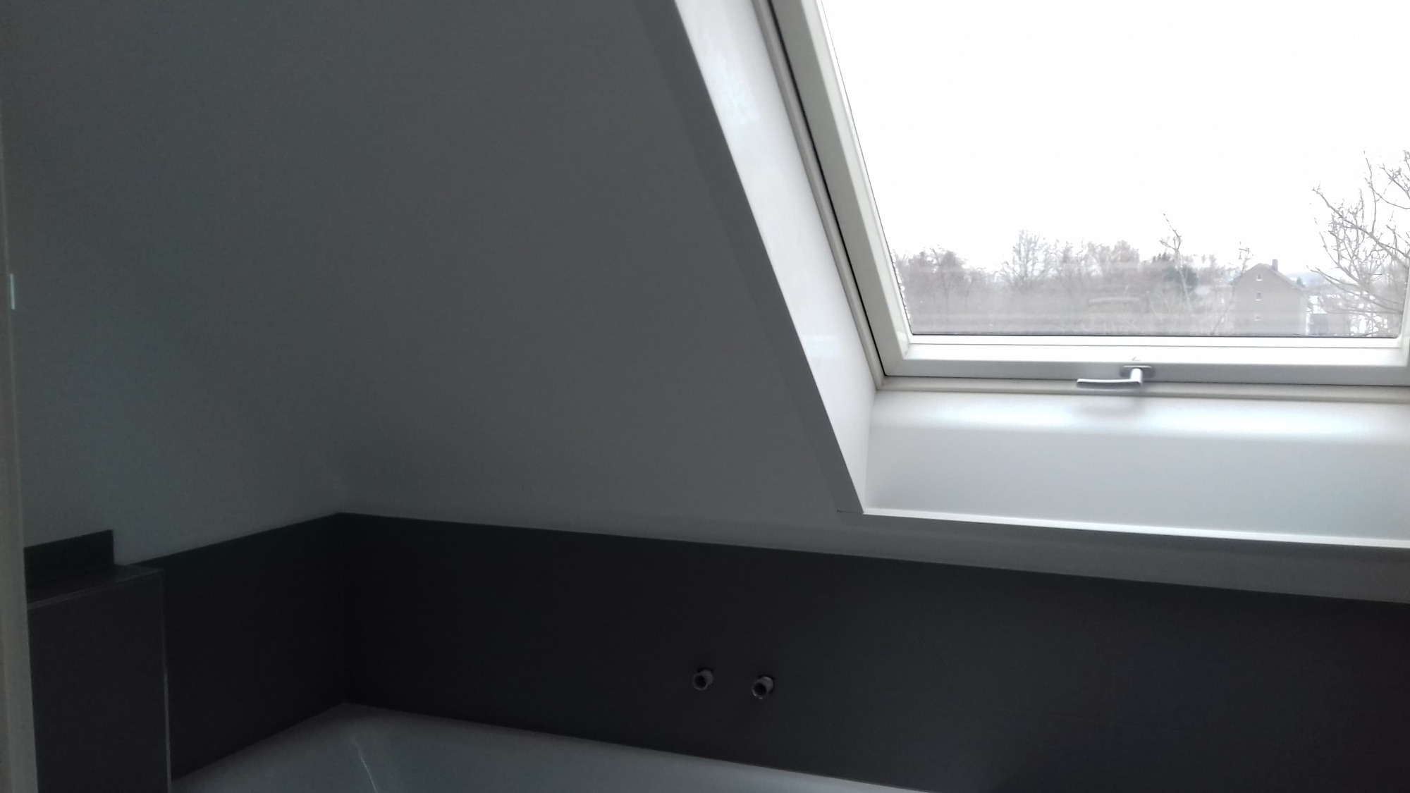 Szare płytki częściowo ułożone na ścianie w łazience na poddaszu, widoczne dwa zawory, okno dachowe z widokiem na zewnątrz.