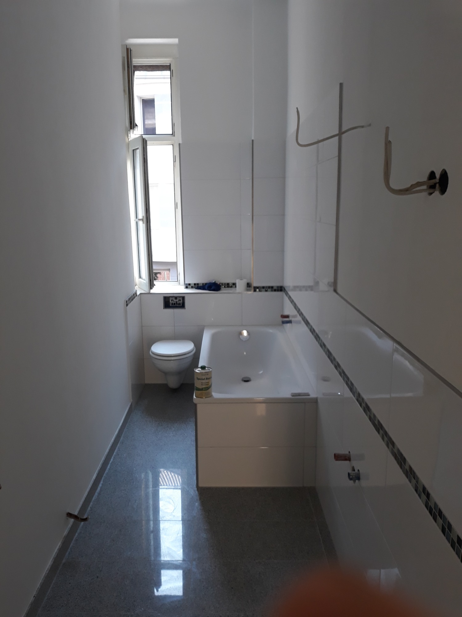 Wąska łazienka w trakcie remontu: białe płytki z dekoracyjnym paskiem, surowy beton na podłodze, otwór okienny z widokiem na budynek, instalacja elektryczna na ścianie, toaleta i wanna w strefie...