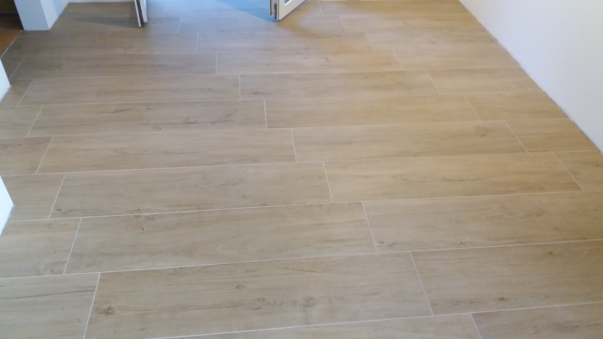 Ułożona podłoga z płytek imitujących drewno w odcieniach beżu i brązu, z widocznymi fugami w kolorze zbliżonym do płytek, prowadząca w kierunku otwartych drzwi.