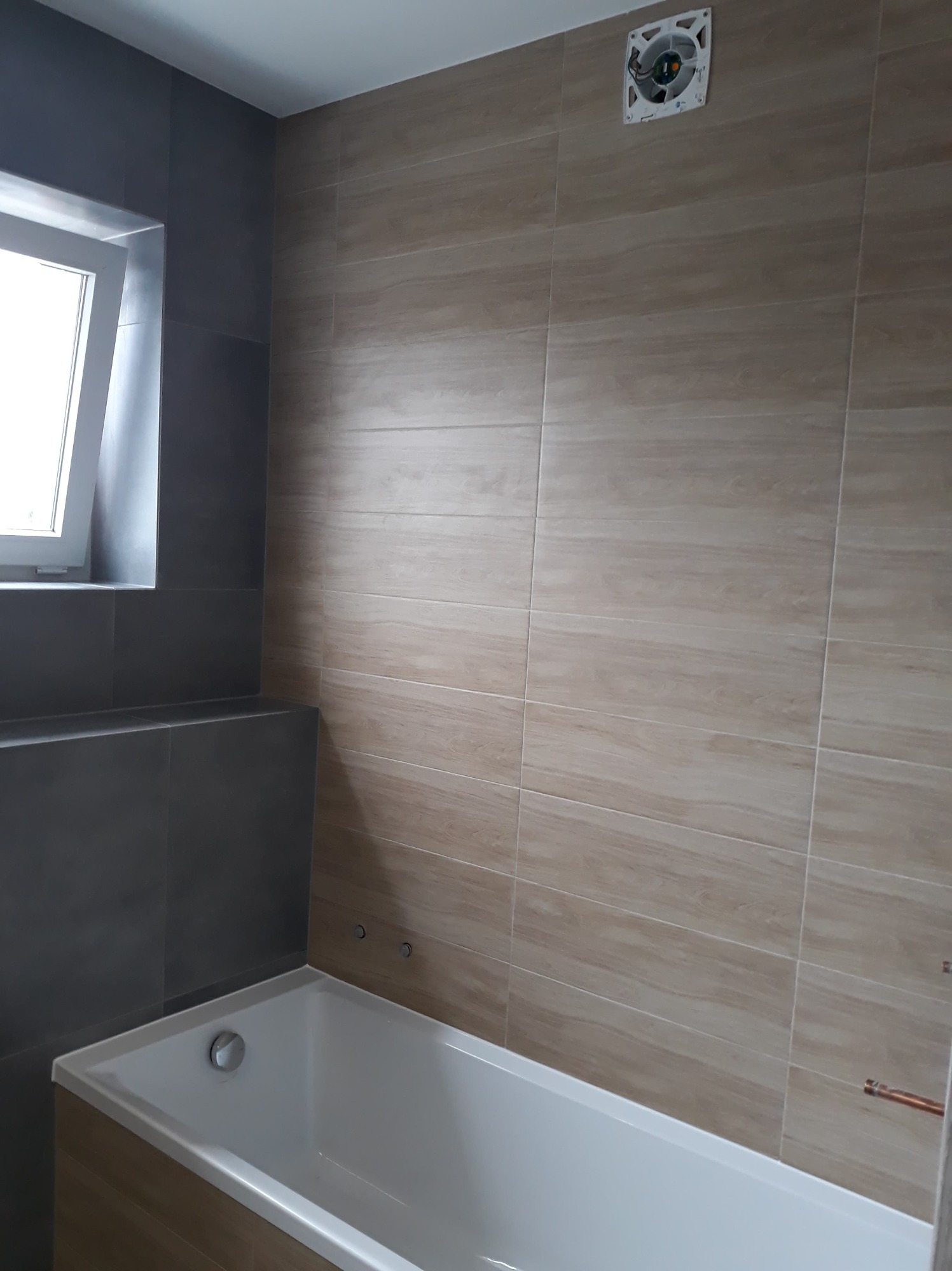 Nowoczesna łazienka z beżowymi płytkami imitującymi drewno na ścianie, białą wanną i szarą zabudową okna. Widoczny wentylator sufitowy.