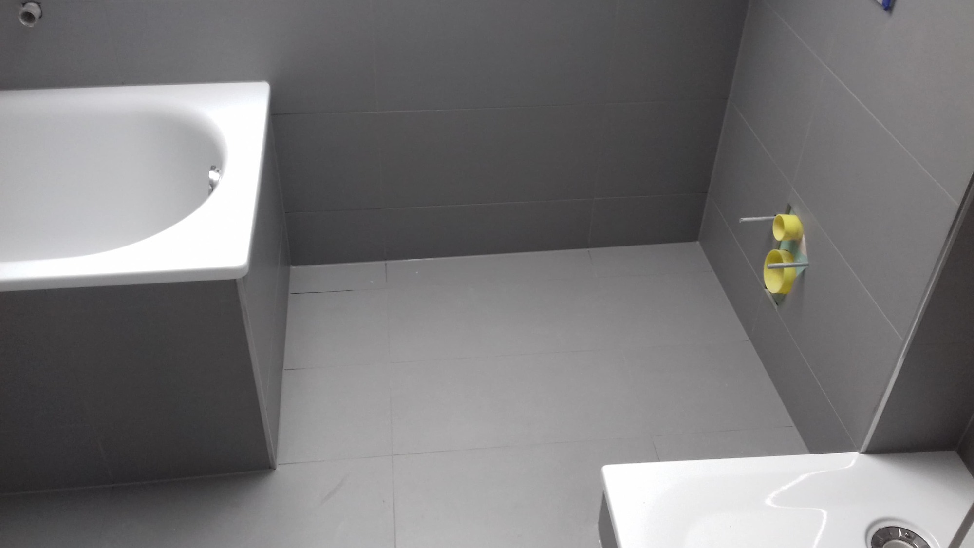 Szara łazienka w trakcie wykańczania, widoczna wanna, umywalka i fragment ściany z rurami zabezpieczonymi żółtymi osłonami, świeżo położone płytki podłogowe i ścienne.