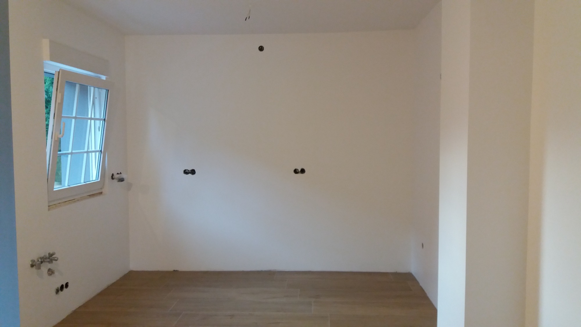 Puste pomieszczenie w trakcie remontu z drewnopodobną podłogą, oknem z białymi ramami i instalacją elektryczną oraz hydrauliczną wystającą ze ścian.