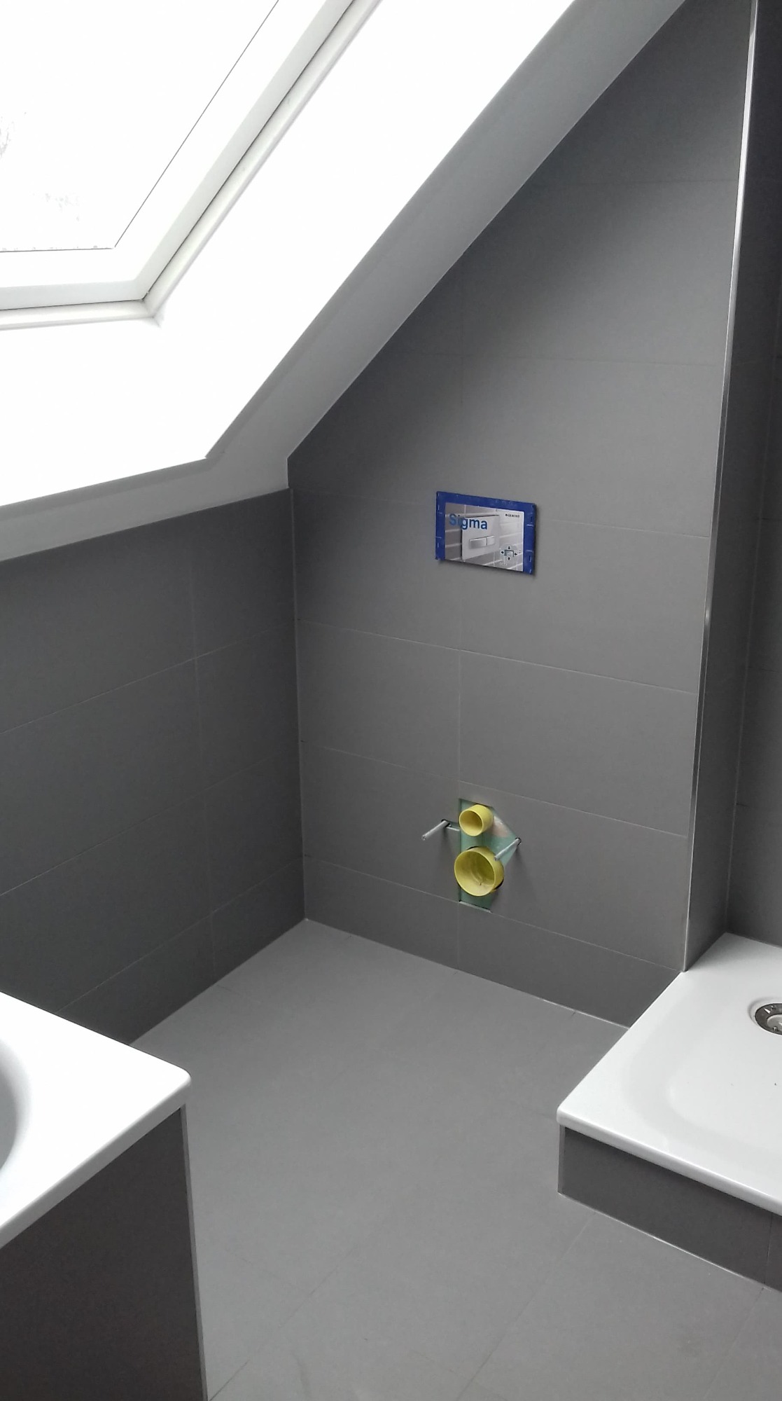 Szara łazienka na poddaszu z oknem dachowym, widoczne płytki ceramiczne, instalacja podtynkowa WC Geberit Sigma i fragment brodzika oraz umywalki.