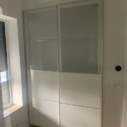 Biała, przesuwna szafa wnękowa z matowego szkła w nowoczesnym wnętrzu, widok z boku okna z parapetem.