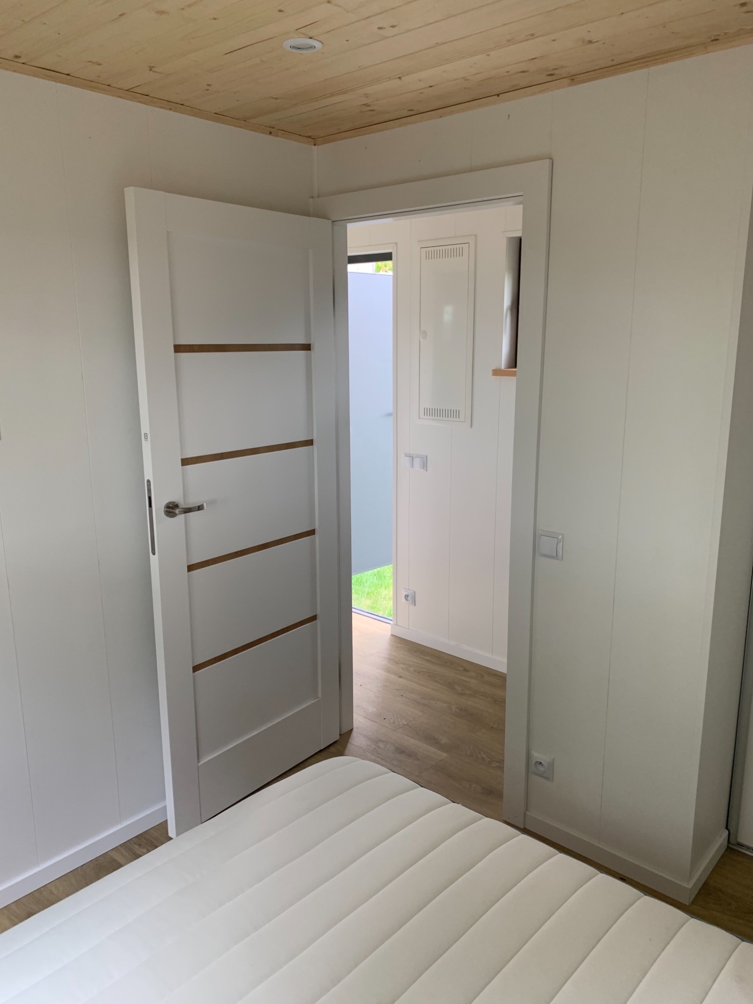 Wnętrze pokoju w jasnych barwach z drewnianą podłogą i sufitem, widoczne białe ściany, białe drzwi z poziomymi drewnianymi wstawkami i fragment białego materaca.