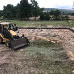 Żółta koparko-ładowarka Caterpillar na placu budowy, obok wykopu pod fundamenty, widoczne drewniane słupki wyznaczające granice, w tle zielone drzewa i słup wysokiego napięcia.