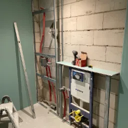 Stelaż podtynkowy Geberit zamontowany na tle ściany z bloczków, widoczne rury instalacyjne, profil metalowy i narzędzia, etap prac w pomieszczeniu z zielonymi ścianami.