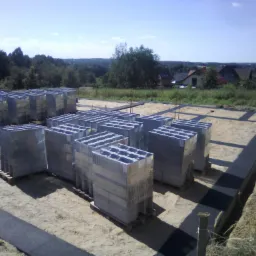 Przygotowany teren pod budowę domu z wykopanymi fundamentami i ułożonymi bloczkami betonowymi na paletach, widoczne czarne pasy izolacji, w tle zabudowa jednorodzinna i zieleń.