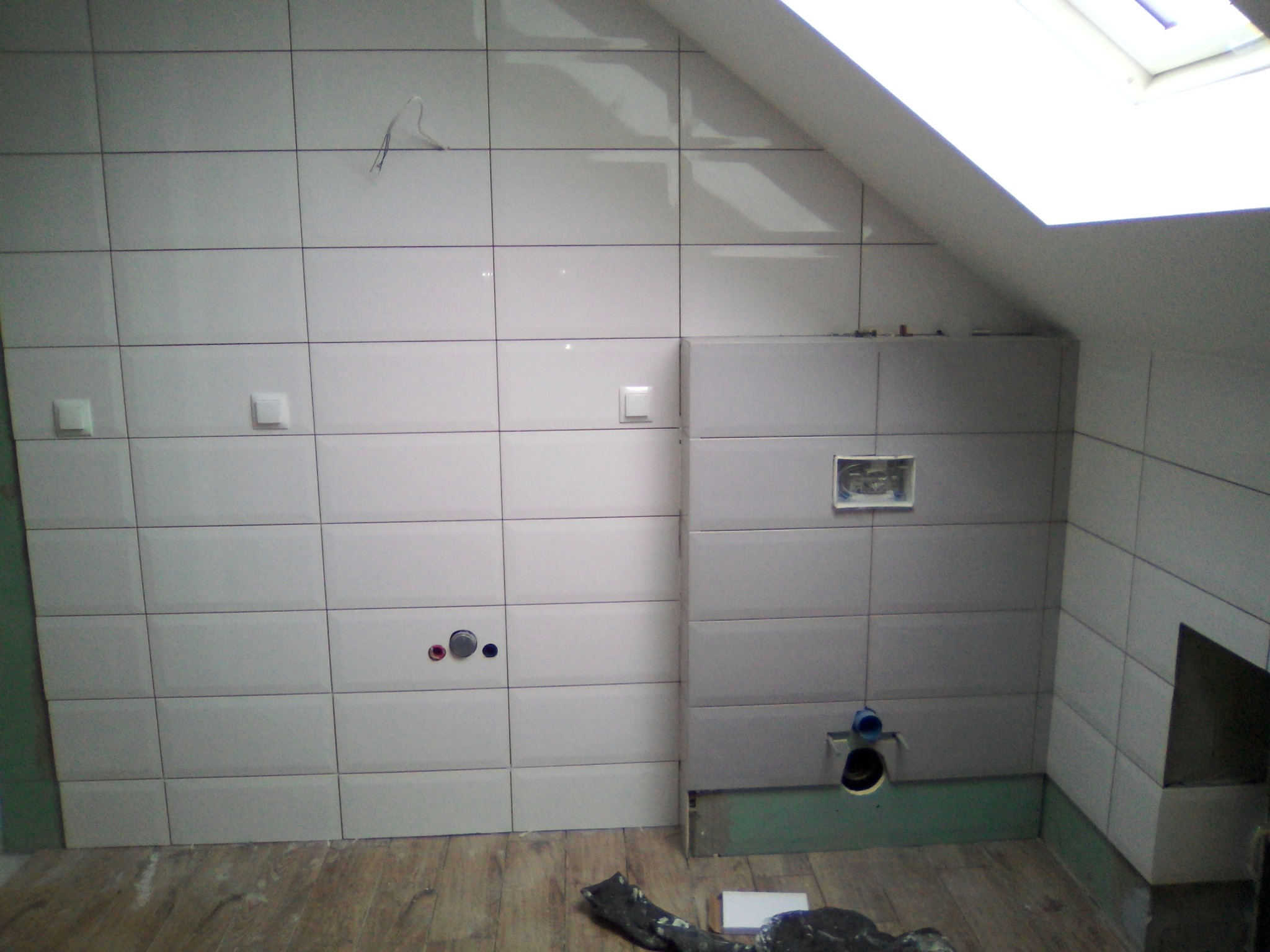 Białe, prostokątne płytki ścienne w trakcie układania w łazience na poddaszu ze świetlikiem, widoczne przyłącza wodne i odpływowe, resztki zaprawy na podłodze.