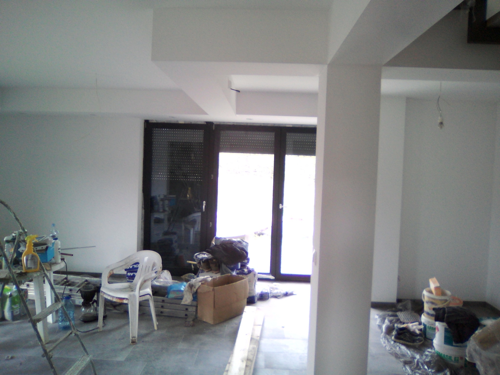 Wnętrze w trakcie wykańczania: białe ściany, surowy beton na podłodze, okno z roletami, drabina, plastikowe krzesło i pojemniki z farbą.