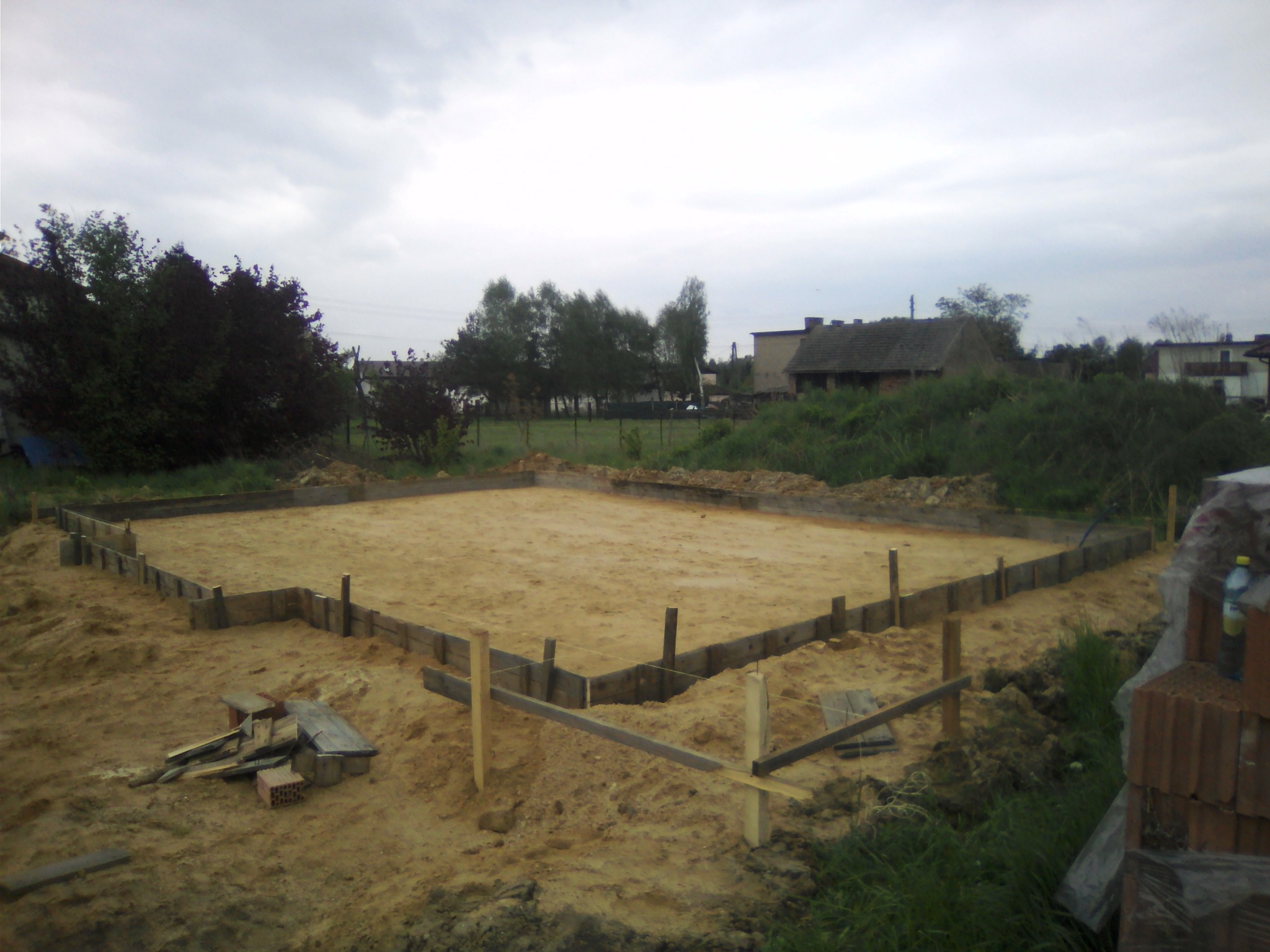 Przygotowany szalunek pod wylewkę fundamentów domu jednorodzinnego na piaszczystym terenie, widoczne drewniane deski i podpory, w tle zarośla i zabudowania.