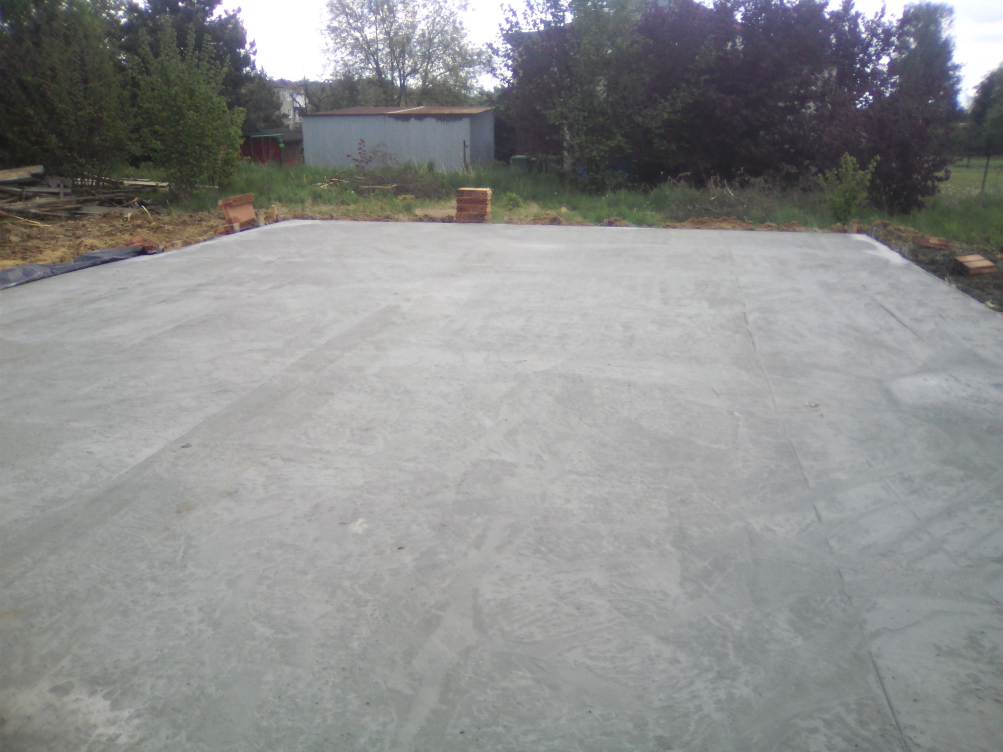 Wylana, szara płyta fundamentowa na przygotowanym terenie, widoczne ułożone cegły na obrzeżach, w tle zielona roślinność i blaszany garaż.