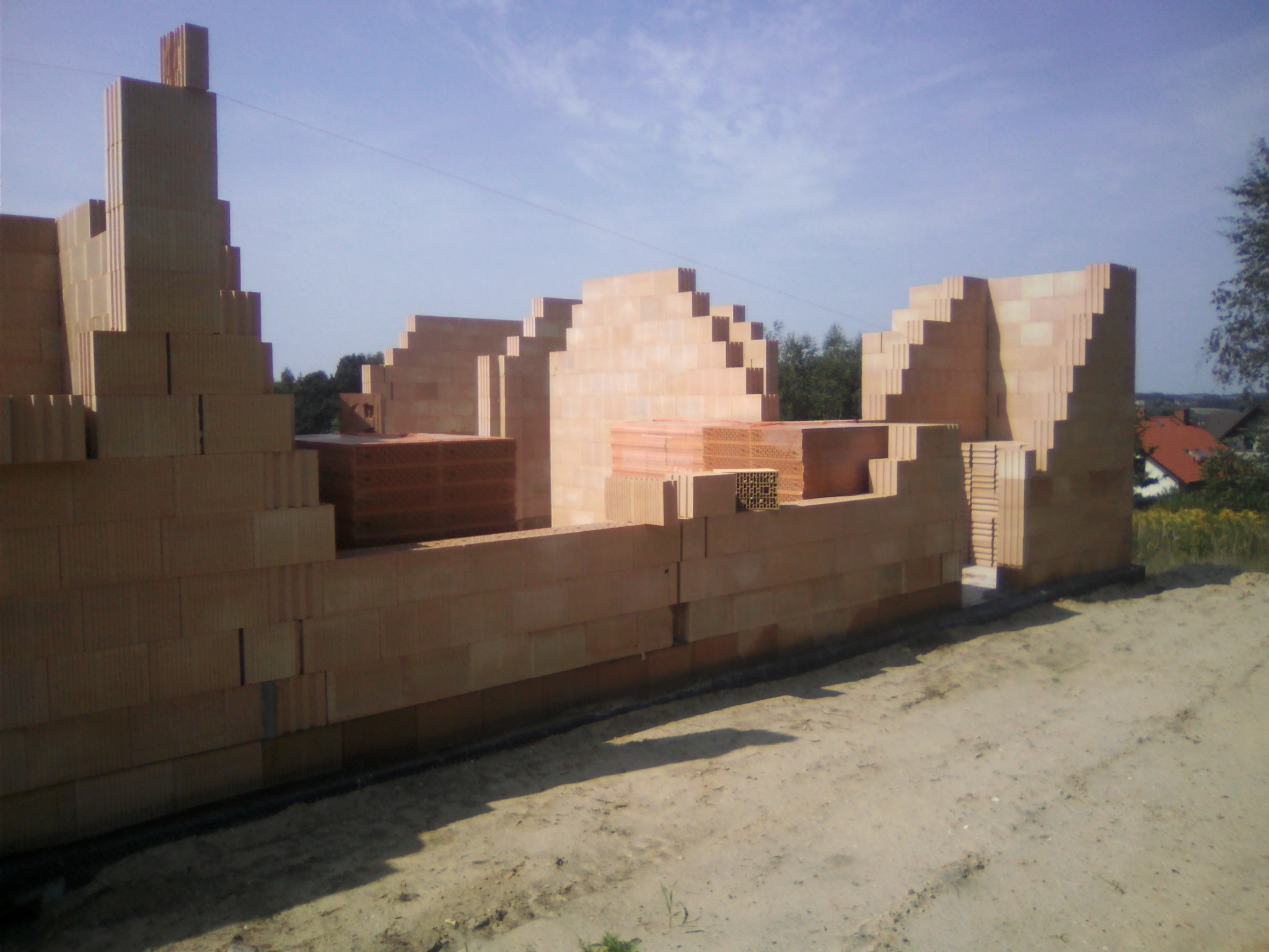 Wznoszone ściany domu z pustaków ceramicznych, widoczne palety z materiałem budowlanym, konstrukcja w trakcie budowy, teren wokół wyrównany.