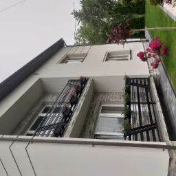 Dwupiętrowy dom z balkonami ozdobionymi czarnymi balustradami i doniczkami z czerwonymi kwiatami, elewacja wykończona kamieniem dekoracyjnym, obok domu trawnik z krzewem rododendronu.