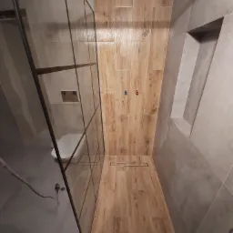 Nowoczesna kabina prysznicowa walk-in z drewnopodobnymi płytkami na ścianie i podłodze, odpływ liniowy, widoczna toaleta w tle, szare płytki na pozostałych ścianach i wnęka w ścianie.