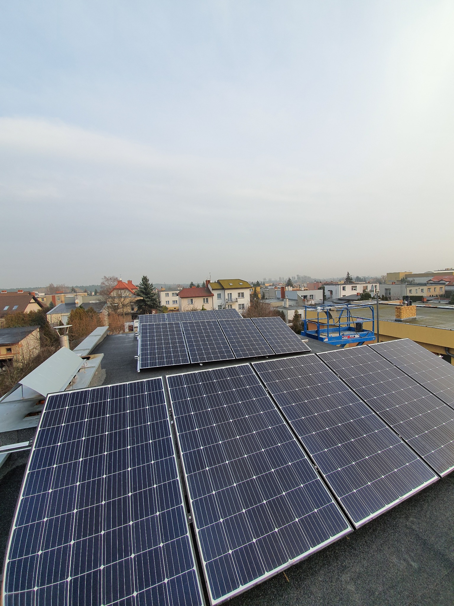 Panel słoneczny na płaskim dachu budynku z widoczną platformą roboczą na tle zabudowy miejskiej w pochmurny dzień.