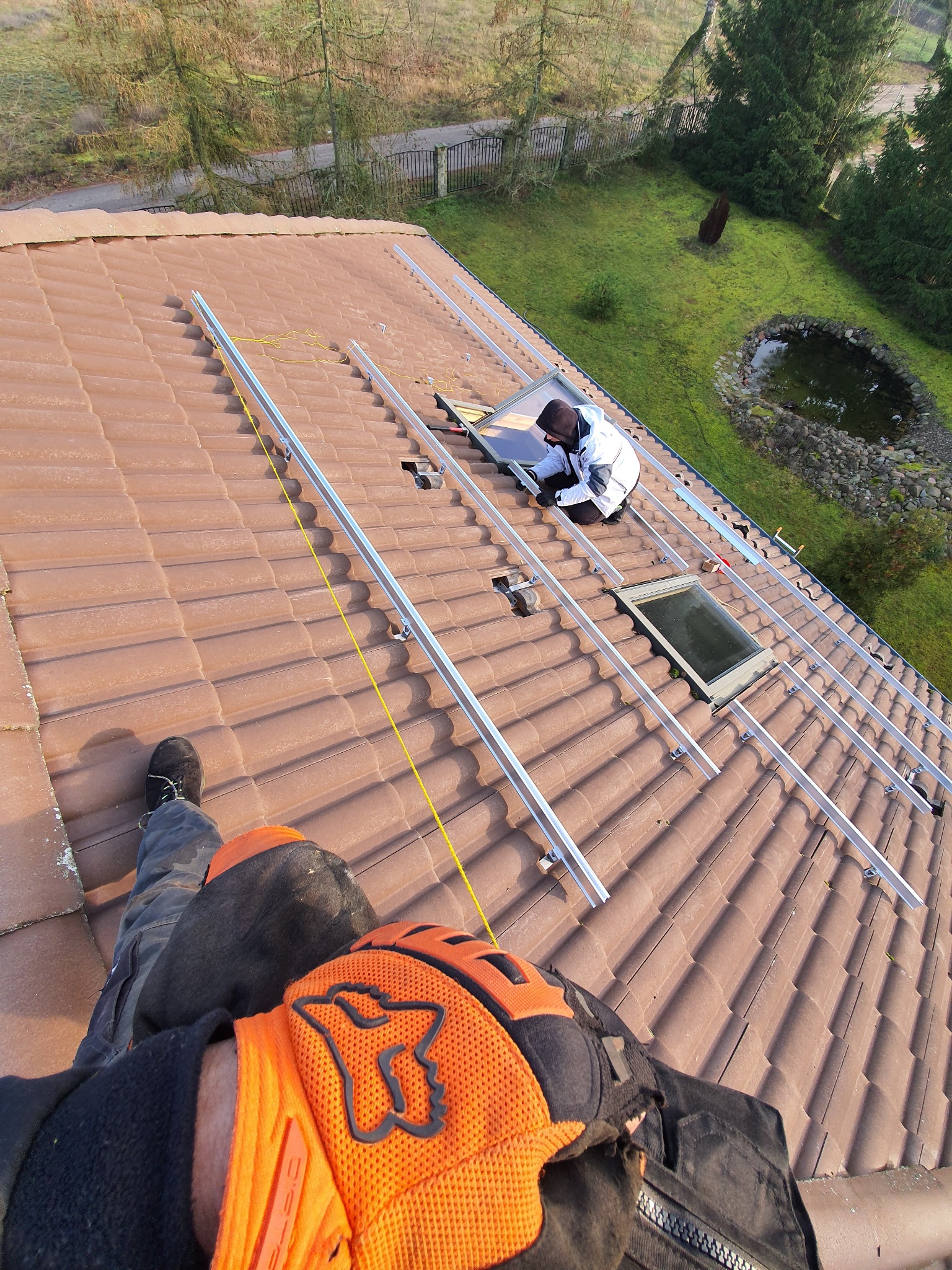 Widok z perspektywy pierwszej osoby na montaż konstrukcji pod panele fotowoltaiczne na brązowym dachu z dachówki ceramicznej, z widocznymi elementami montażowymi, stopą montera w czarnym bucie...