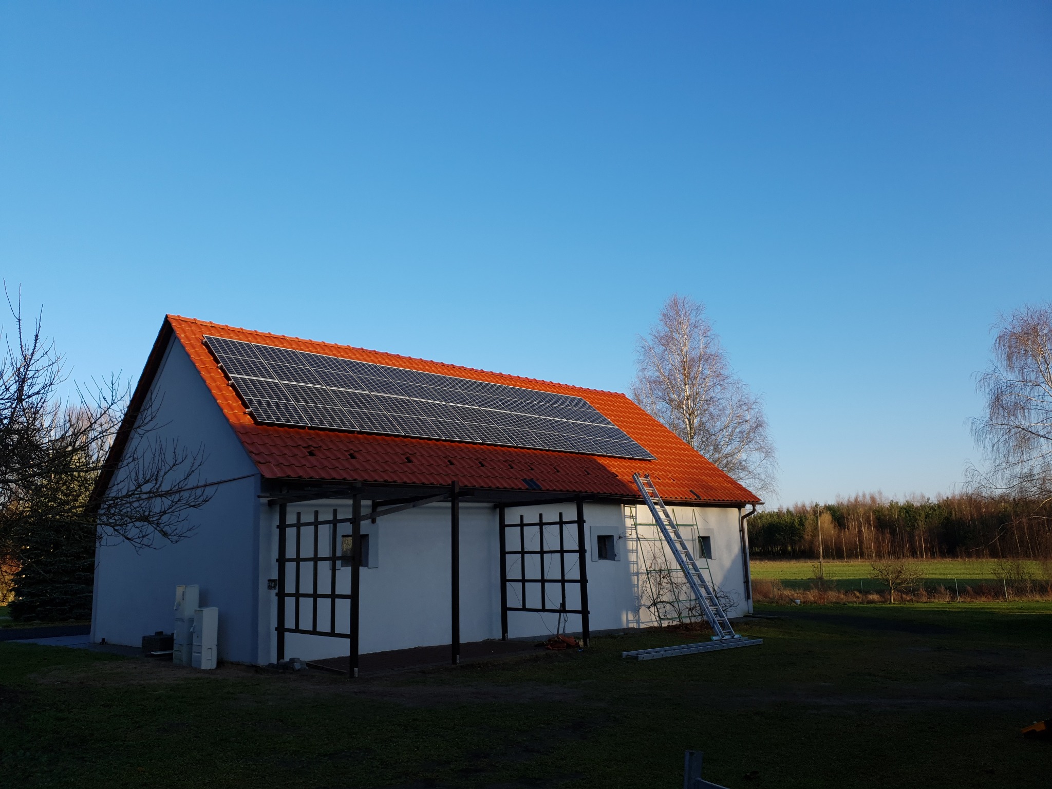 Dom jednorodzinny z panelami słonecznymi na czerwonym dachu w słoneczny dzień w okolicach Poznania. Rozstawiona drabina oparta o ścianę budynku sugeruje trwające prace instalacyjne.
