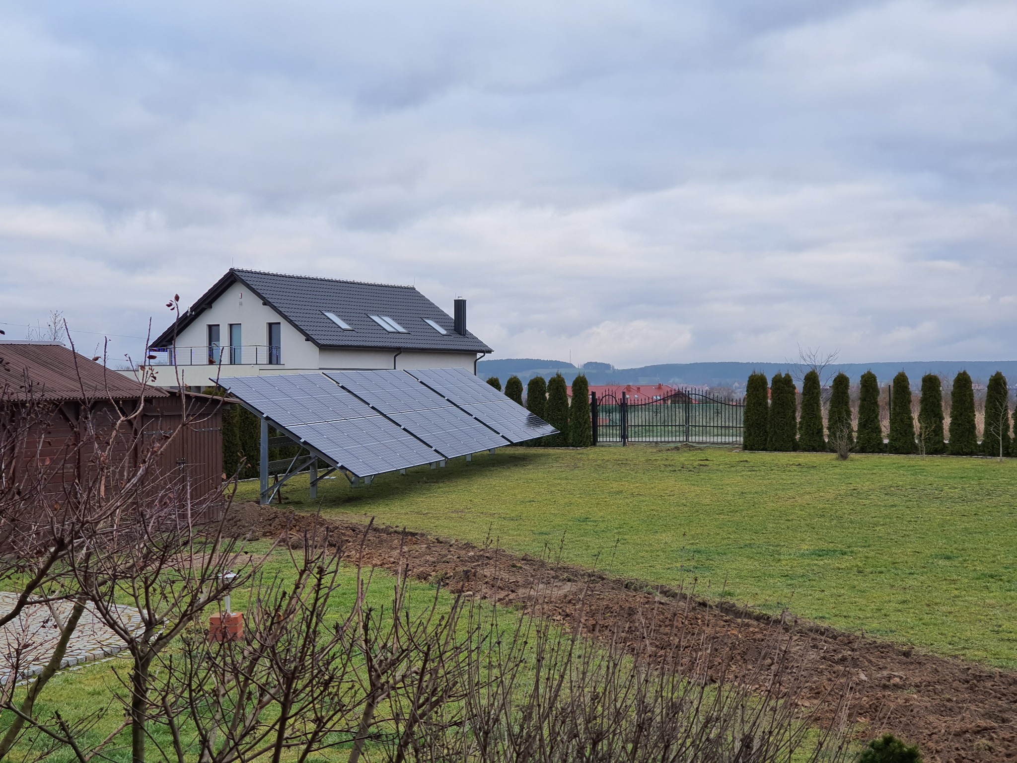 Instalacja paneli słonecznych na gruncie przed domem jednorodzinnym z widocznymi śladami prac ziemnych, w tle rząd tuj i ogrodzenie.