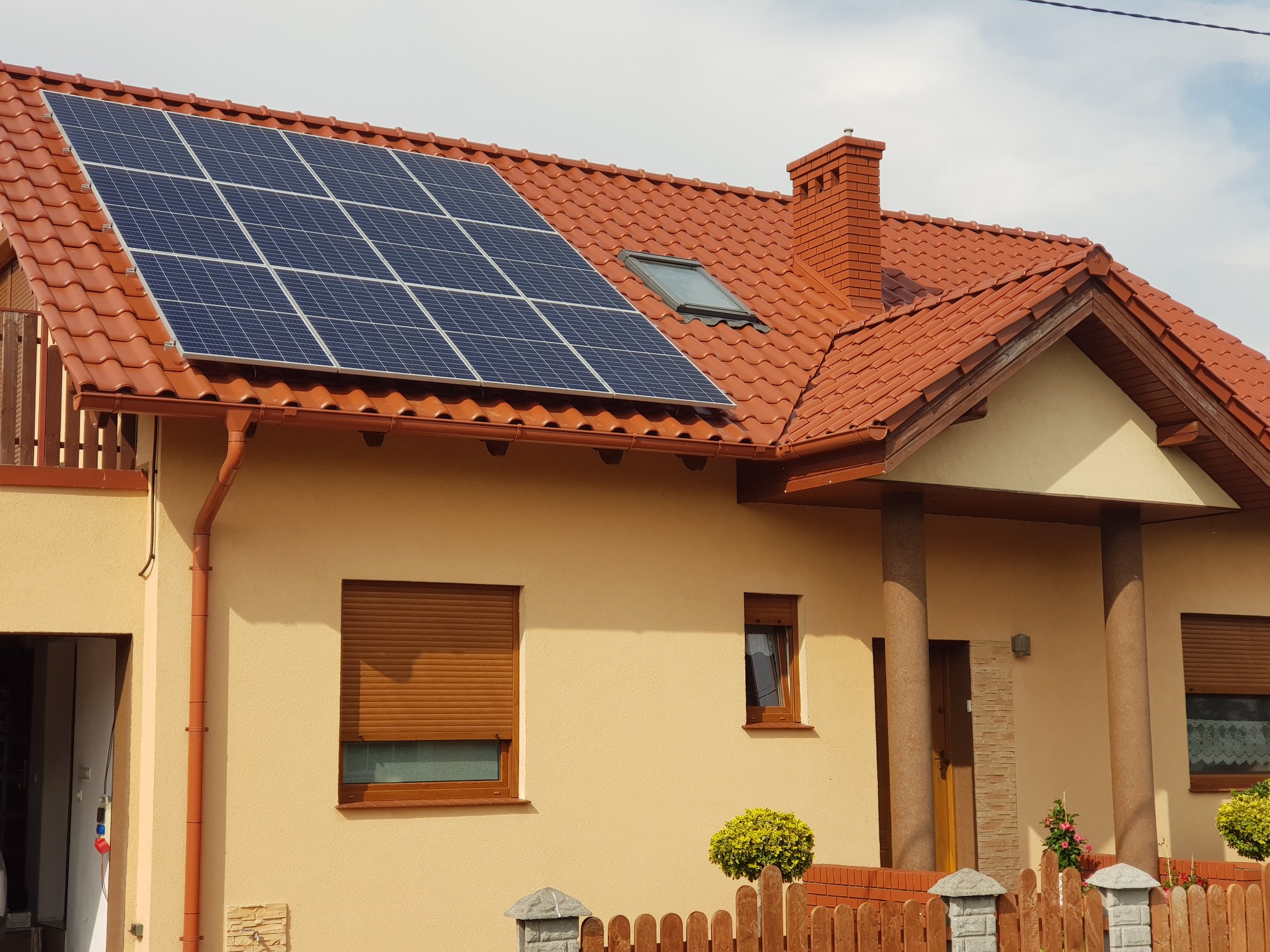 Dom jednorodzinny z panelami słonecznymi na czerwonym dachu i brązowymi roletami w oknach.