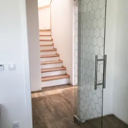 Szklane drzwi z geometrycznym wzorem plastra miodu, prowadzące do klatki schodowej z drewnianymi stopniami, widoczne chromowane okucia i uchwyt.