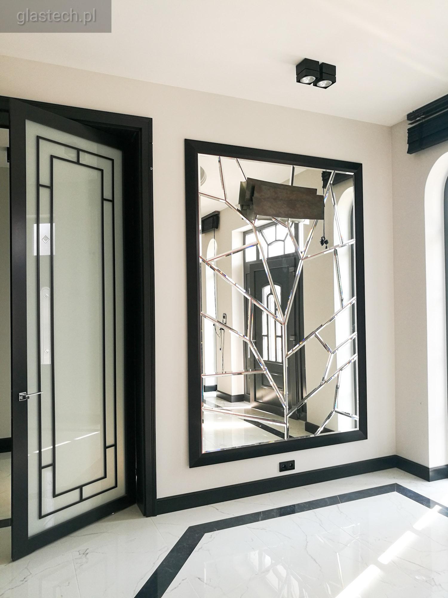 Nowoczesne lustro dekoracyjne z geometrycznym podziałem tafli w czarnej ramie, odbijające drzwi z przeszkleniami w holu z marmurową posadzką.