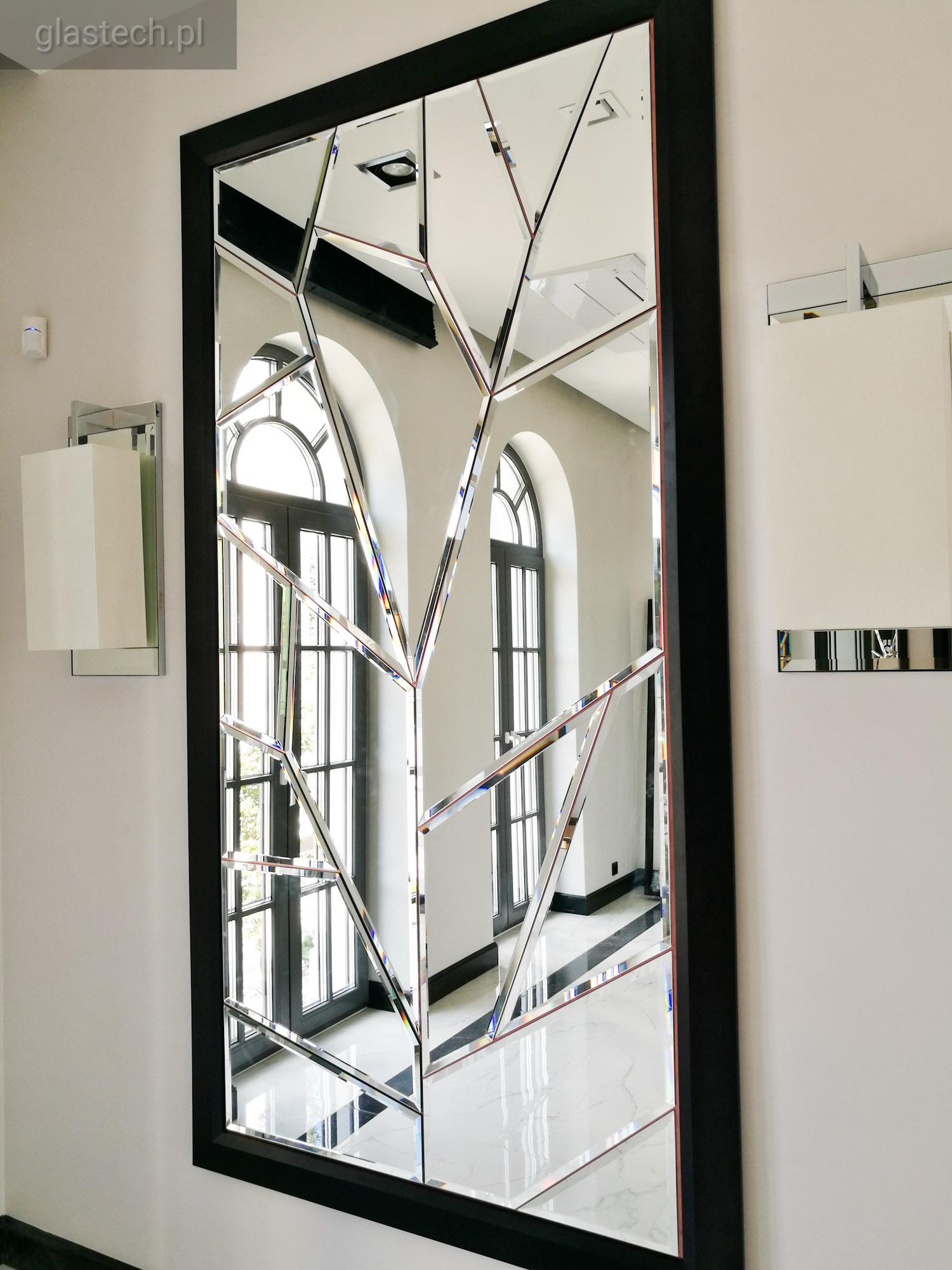 Duże lustro dekoracyjne z czarną ramą, składające się z nieregularnych tafli, odbijające wnętrze z wysokimi oknami łukowymi i marmurową podłogą. Na ścianie obok lustra zamontowane nowoczesne lampy.