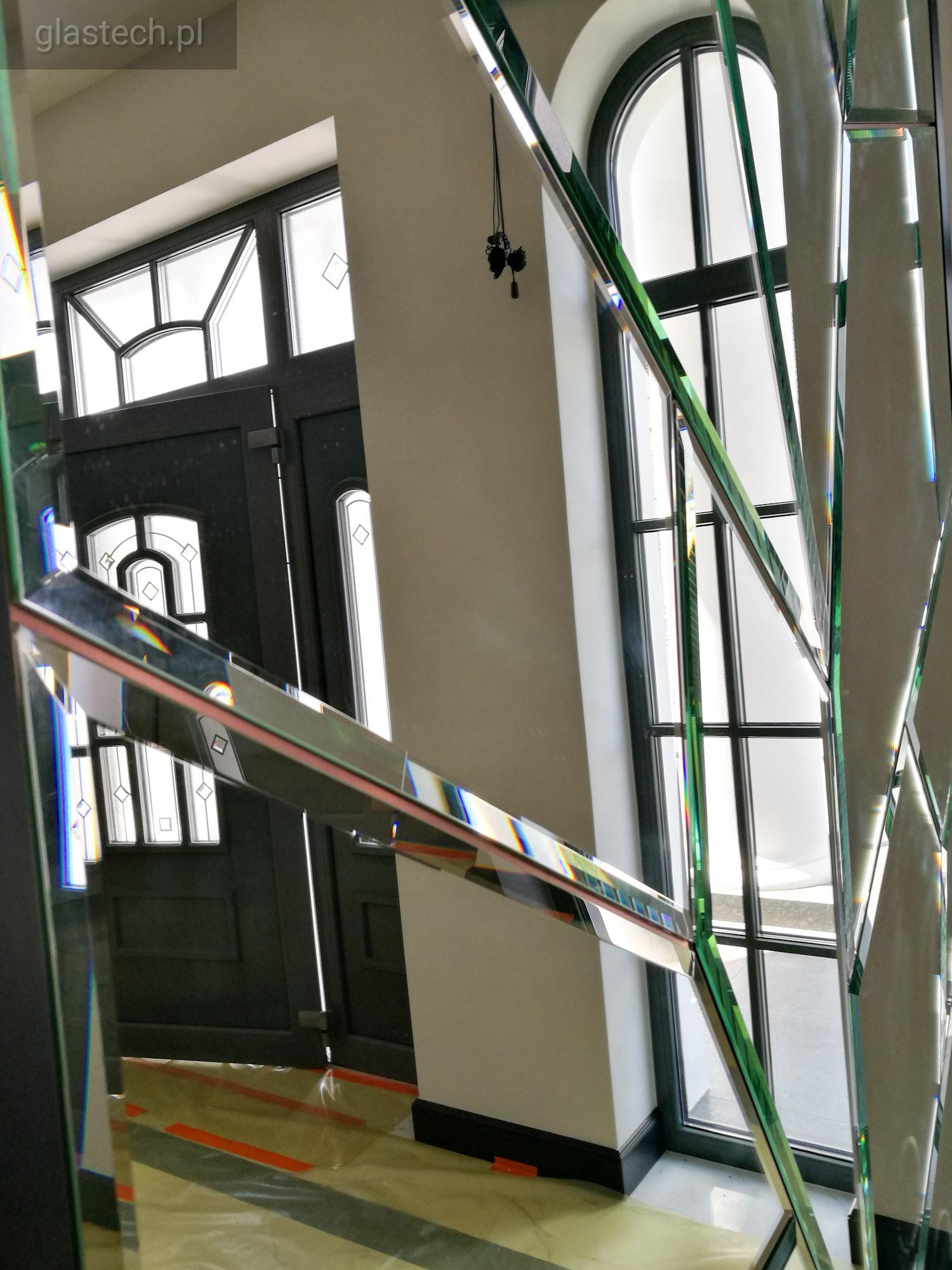 Abstrakcyjna kompozycja z tafli szkła, tworząca iluzję przestrzeni i odbijająca fragmenty drzwi wejściowych oraz okna łukowego w tle.