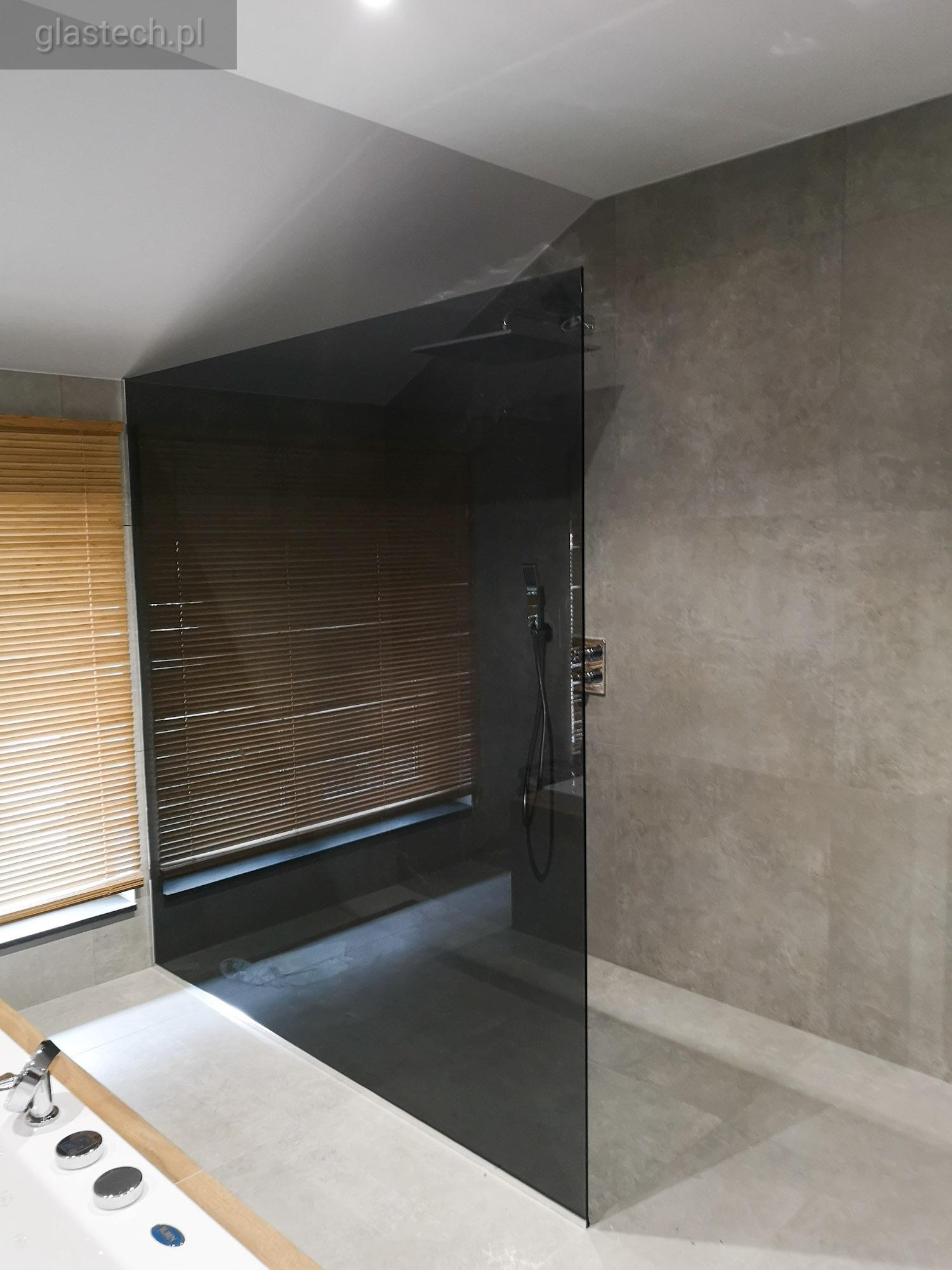 Nowoczesna kabina prysznicowa z przyciemnianego szkła, widoczna bateria i słuchawka prysznicowa, szare płytki na ścianie, żaluzje drewniane w tle.