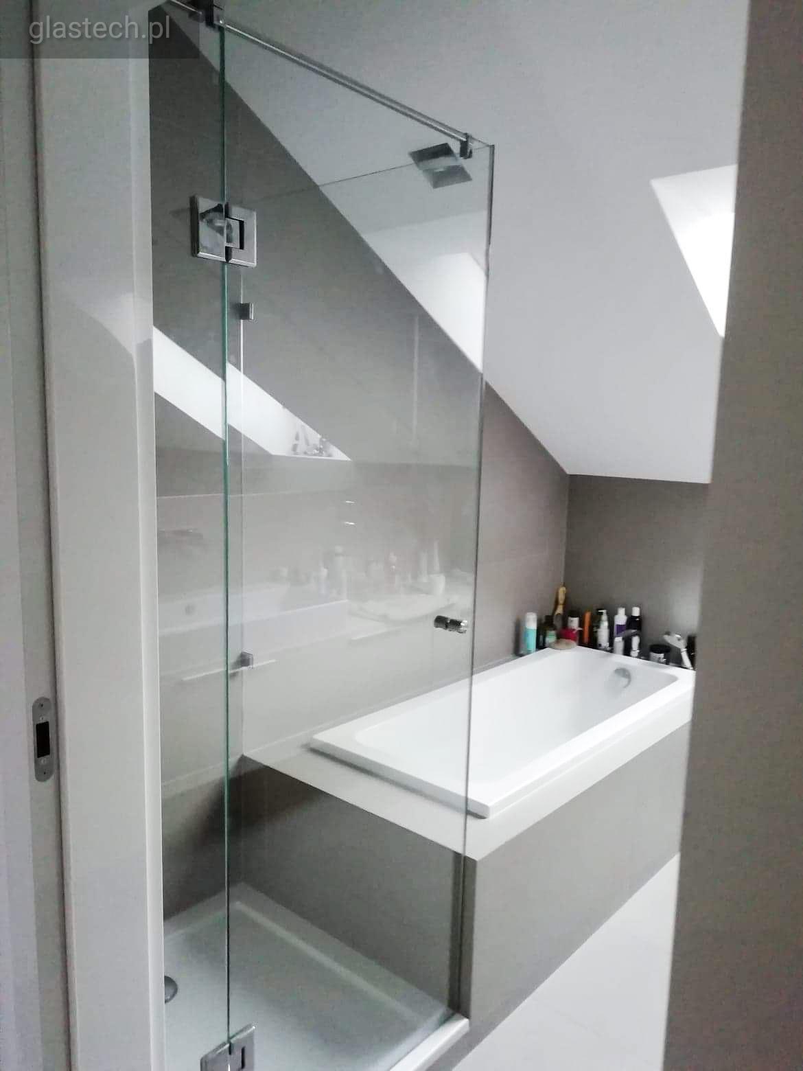 Szklana kabina prysznicowa z chromowanymi zawiasami w łazience na poddaszu, widoczna wanna obudowana szarą płytą.