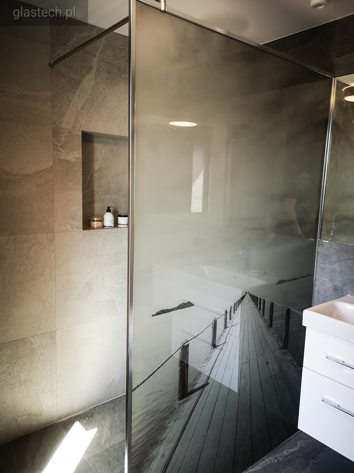 Szklana kabina prysznicowa z motywem długiego drewnianego mola nad wodą, widoczna w tle biała umywalka z szafką, na ścianie wnęka z kosmetykami.
