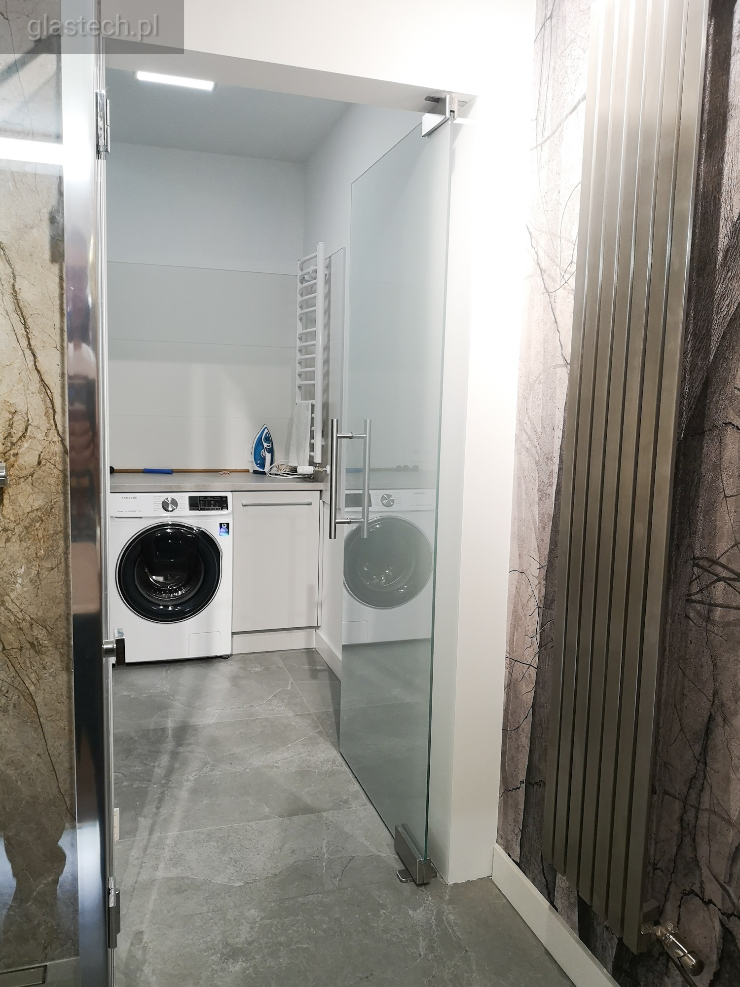 Nowoczesne szklane drzwi wahadłowe z chromowanymi uchwytami i zawiasami, oddzielające łazienkę z pralkami i grzejnikiem dekoracyjnym na ścianie z motywem drzewa.