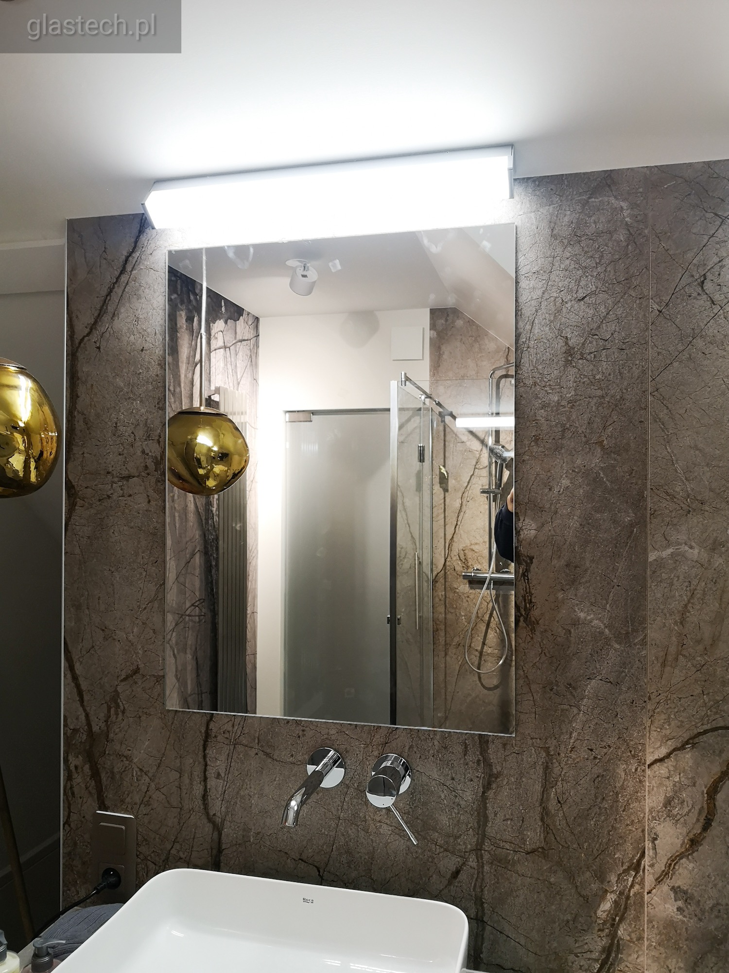 Prostokątne lustro z podświetleniem LED nad umywalką w łazience wykończonej płytkami imitującymi marmur. W odbiciu widoczna kabina prysznicowa ze szklanymi drzwiami.