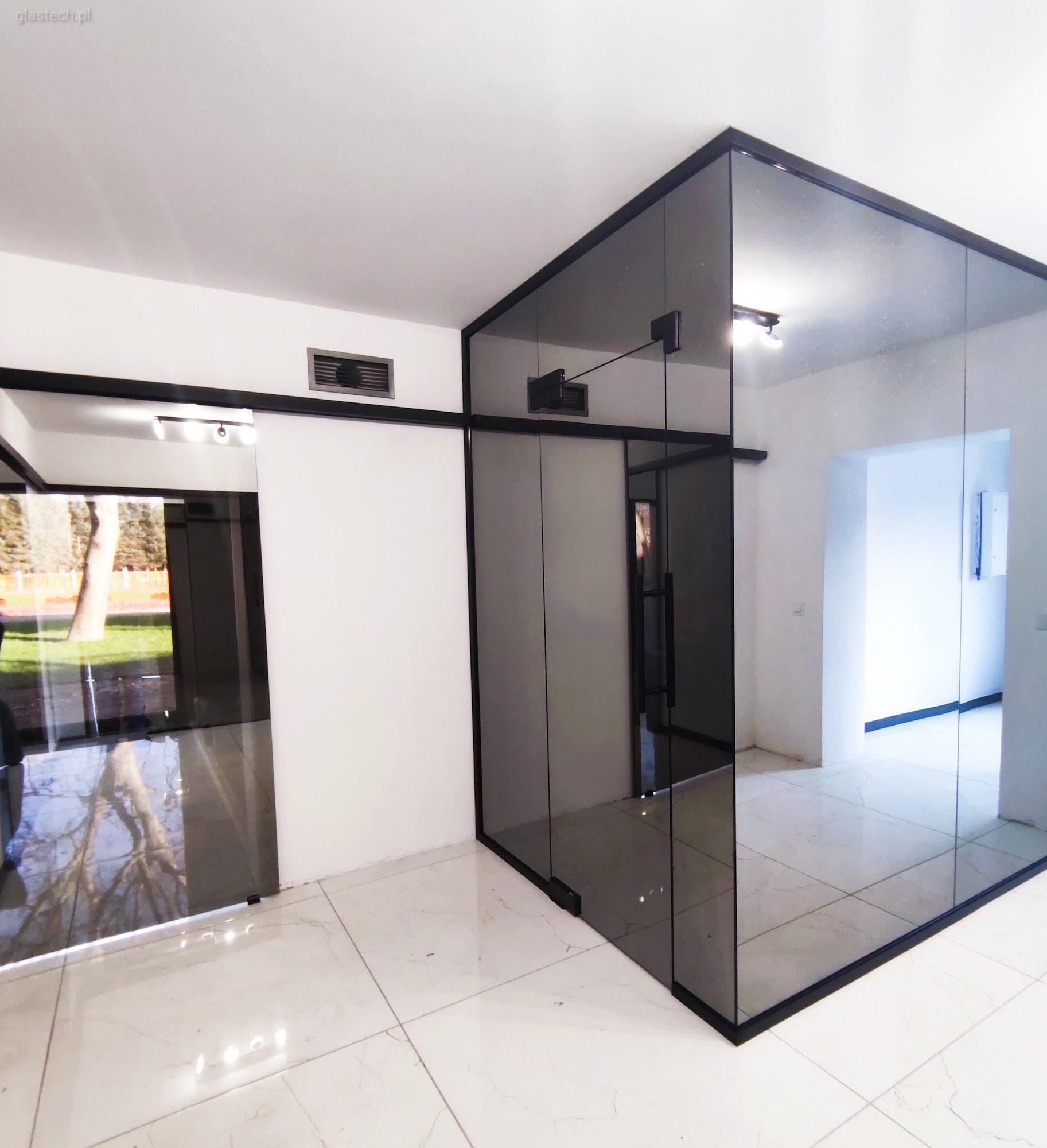 Nowoczesna szklana ścianka działowa z przesuwnymi drzwiami w biurze, czarne ramy, widoczne odbicie otoczenia.