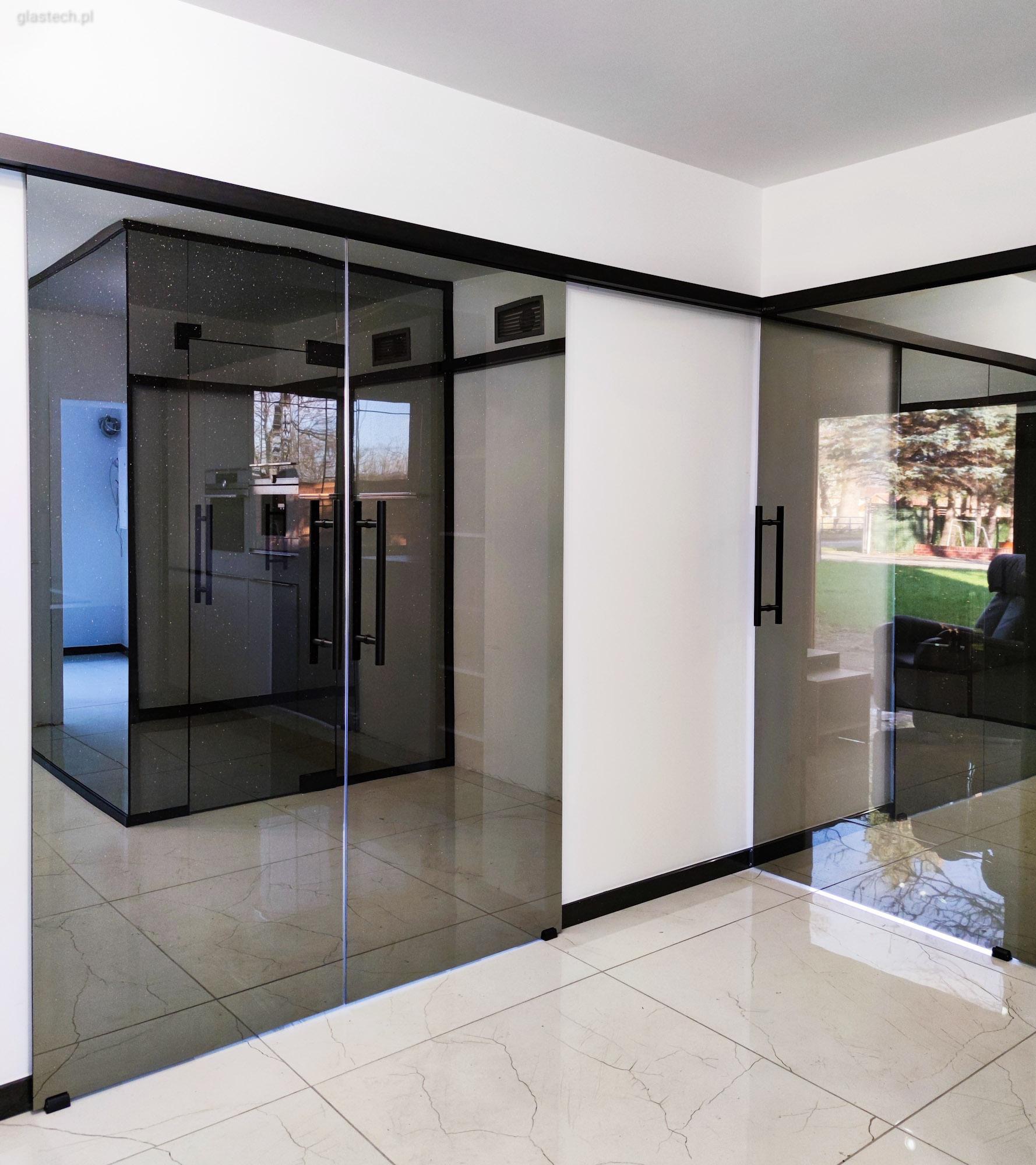 Szklane drzwi przesuwne z czarnymi ramami i uchwytami tworzące nowoczesne przejście między pomieszczeniami, widoczne refleksy na podłodze z płytek.