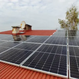 Panele słoneczne zamontowane na czerwonym, blaszanym dachu domu jednorodzinnego, widoczny komin z anteną satelitarną i drzewo w tle.
