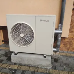 Zewnętrzna jednostka pompy ciepła marki SPRSUN zamontowana na betonowym fundamencie przy ścianie budynku z widoczną rurą odprowadzającą skropliny owiniętą taśmą izolacyjną.