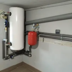 Biały bojler i czerwony zbiornik wyrównawczy zamontowane w kotłowni, widoczne rury w izolacji, manometr i elektroniczny termometr na ścianie.