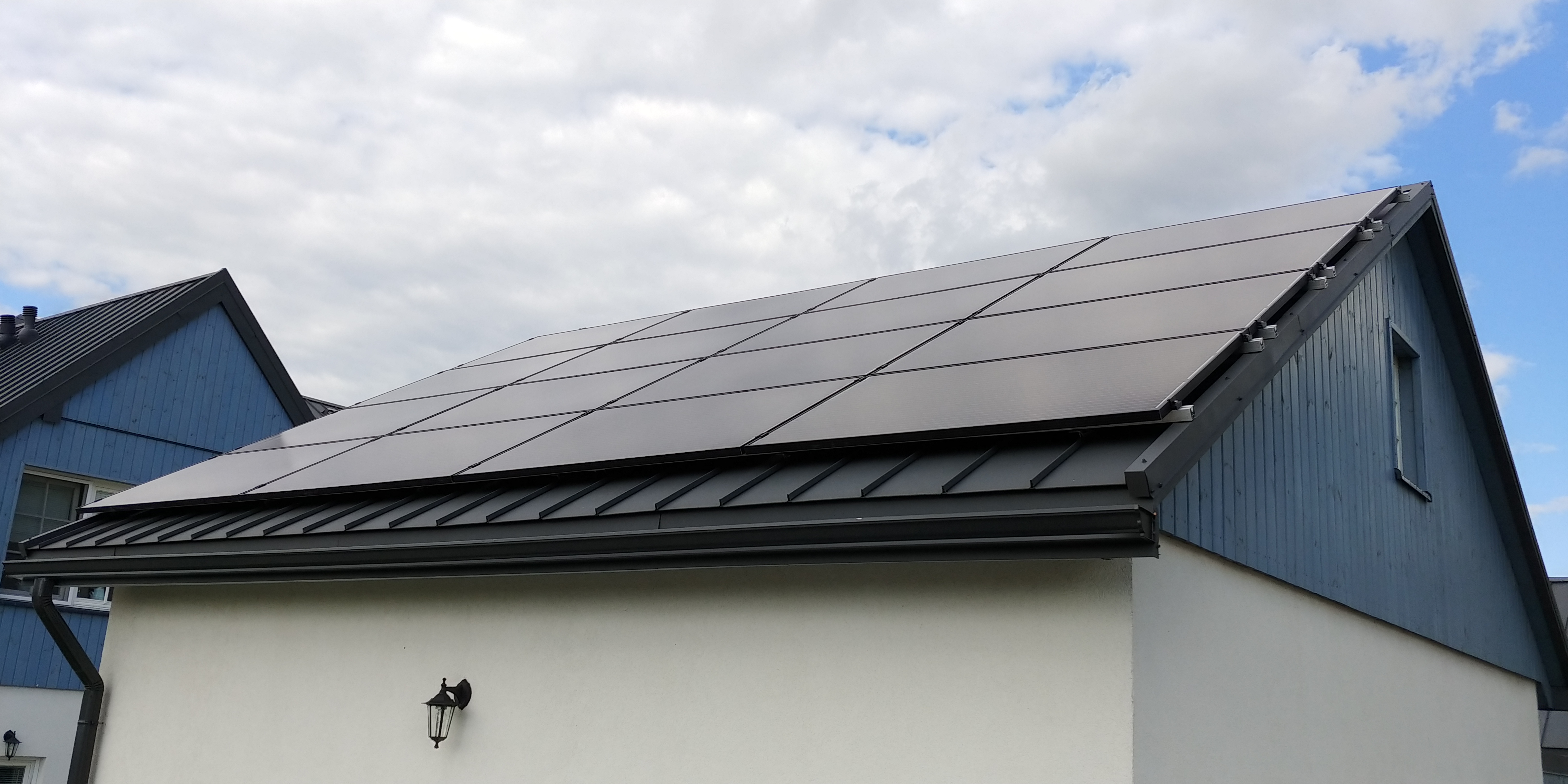 Panele słoneczne na dachu budynku z szarą blachodachówką i białą elewacją, widok z dołu w słoneczny dzień.