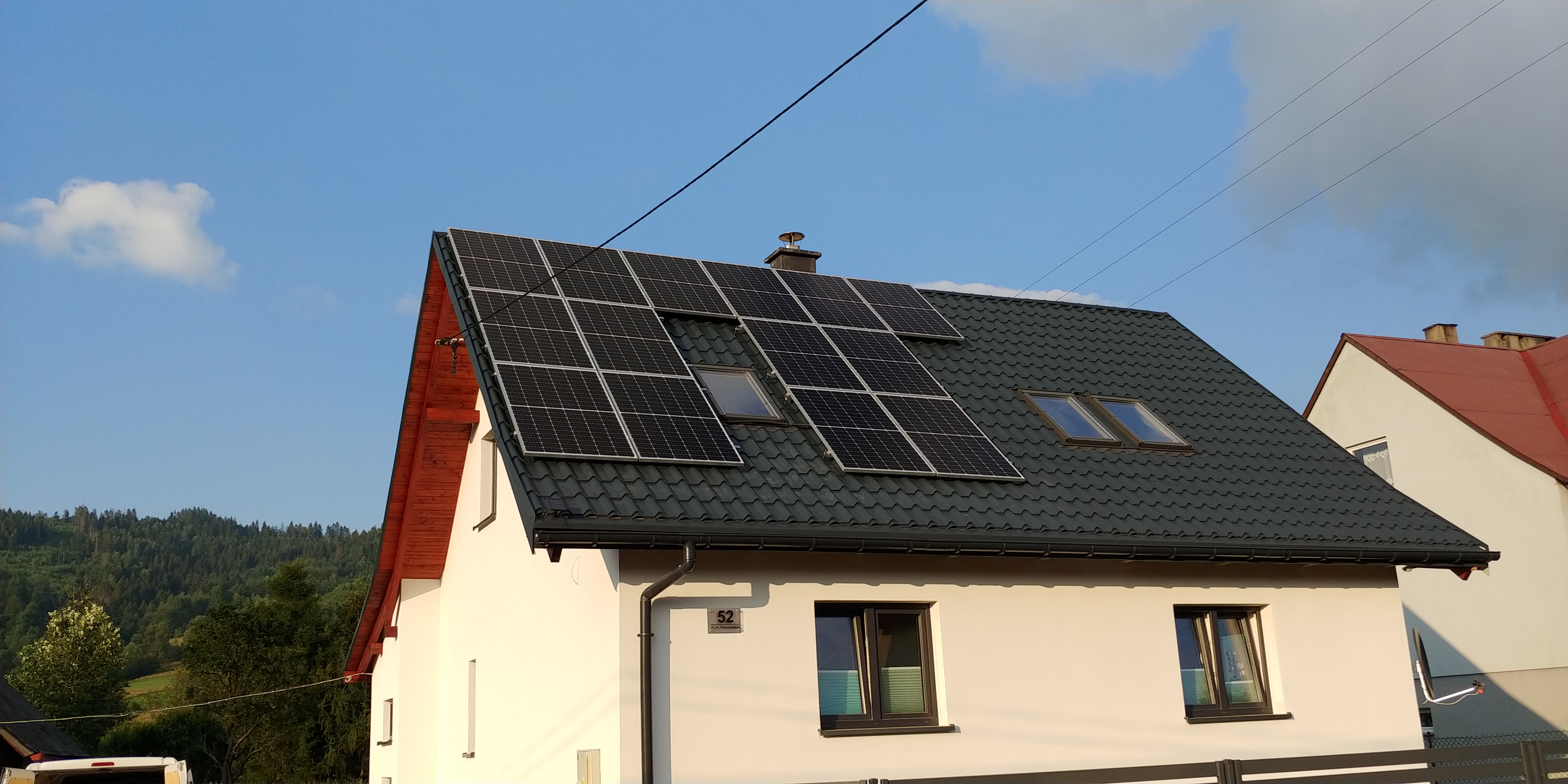 Dom jednorodzinny z panelami słonecznymi na dachu, widok z dołu na elewację i część dachu, w tle zalesione wzgórze.