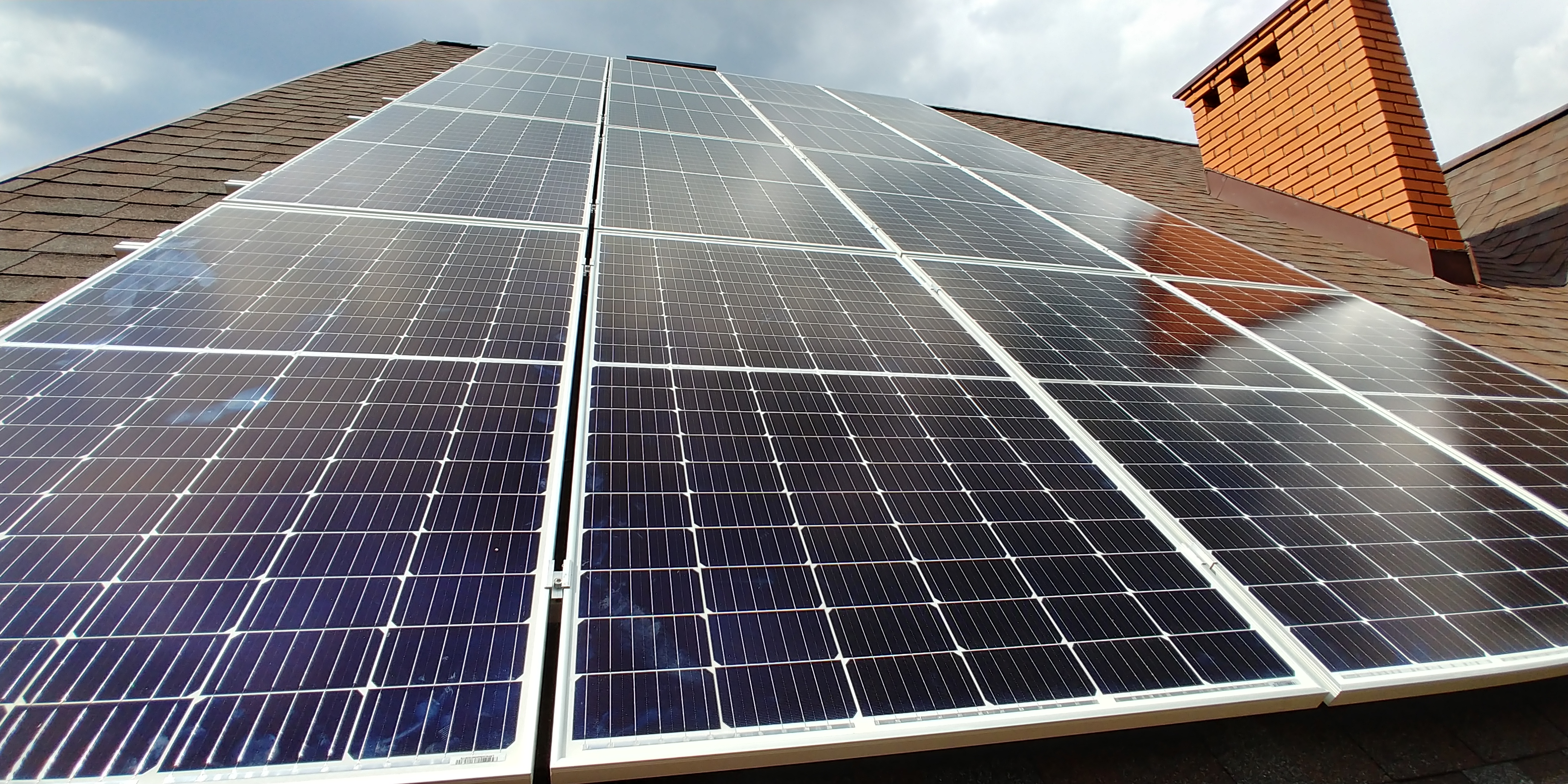 Panele słoneczne zamontowane na dachu z brązowej dachówki, widoczny komin z cegły w tle, pochmurne niebo.