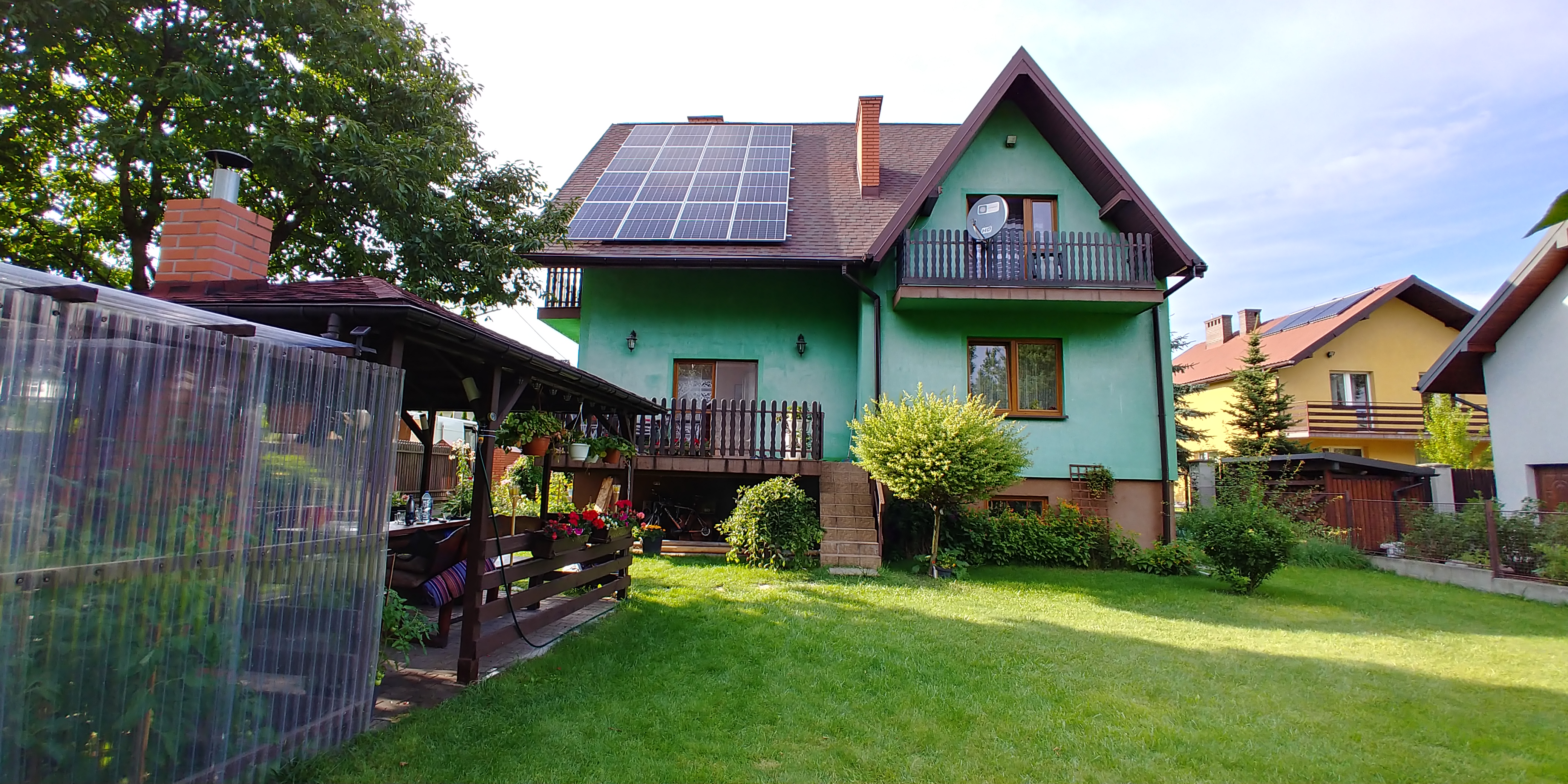 Zielony dom jednorodzinny z brązowym dachem, na którym zamontowane są panele fotowoltaiczne; widok z ogrodu, w tle altana z przezroczystymi ścianami i inne domy.
