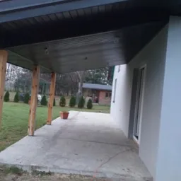 Zadaszenie tarasu z ciemną podbitką, widoczne drewniane słupy i betonowa podłoga, w tle trawnik z tujami i budynek gospodarczy.