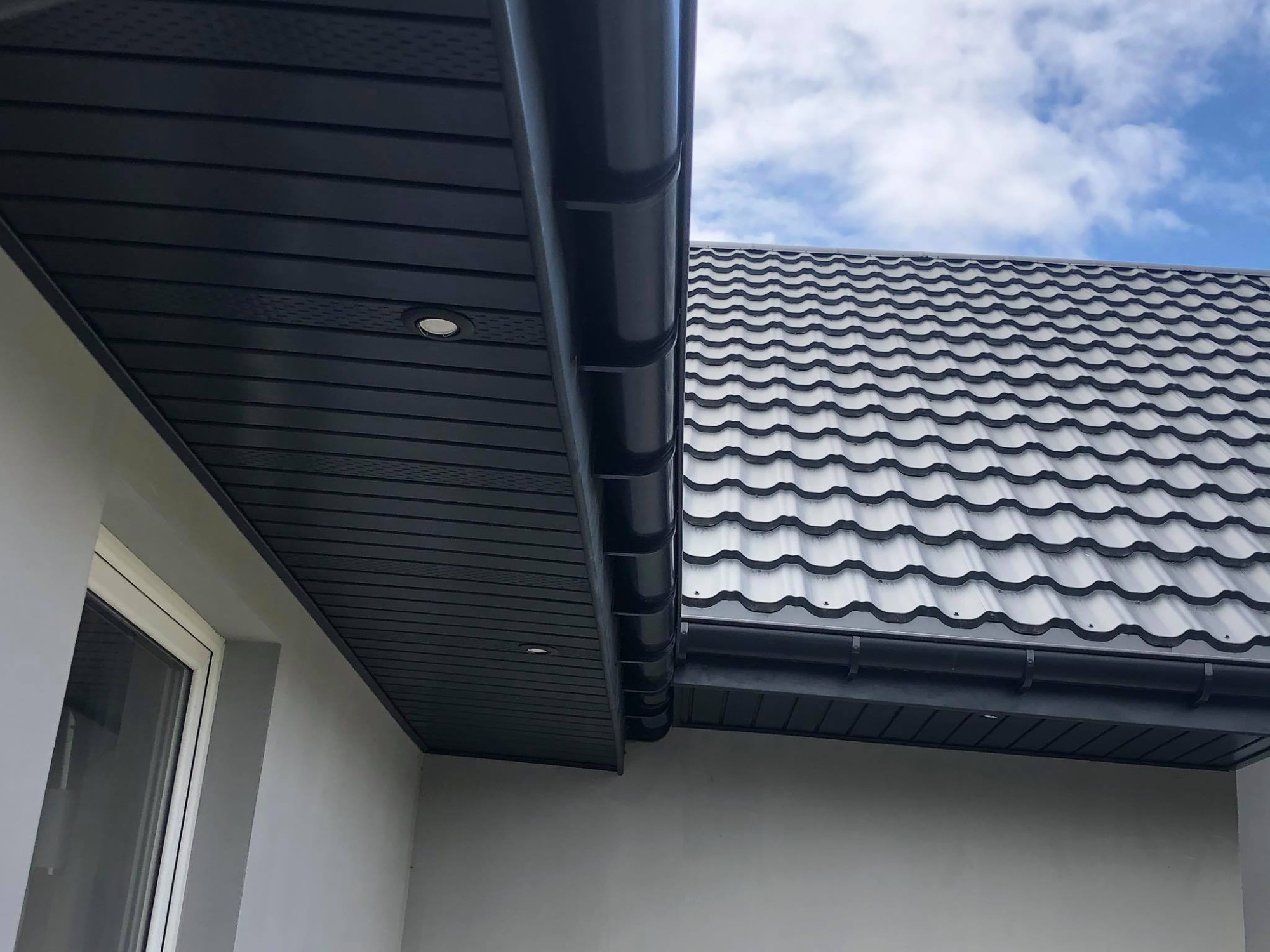 Nowoczesny, grafitowy dach z blachodachówki, widoczne orynnowanie i podbitka z oświetleniem punktowym, błękitne niebo w tle.